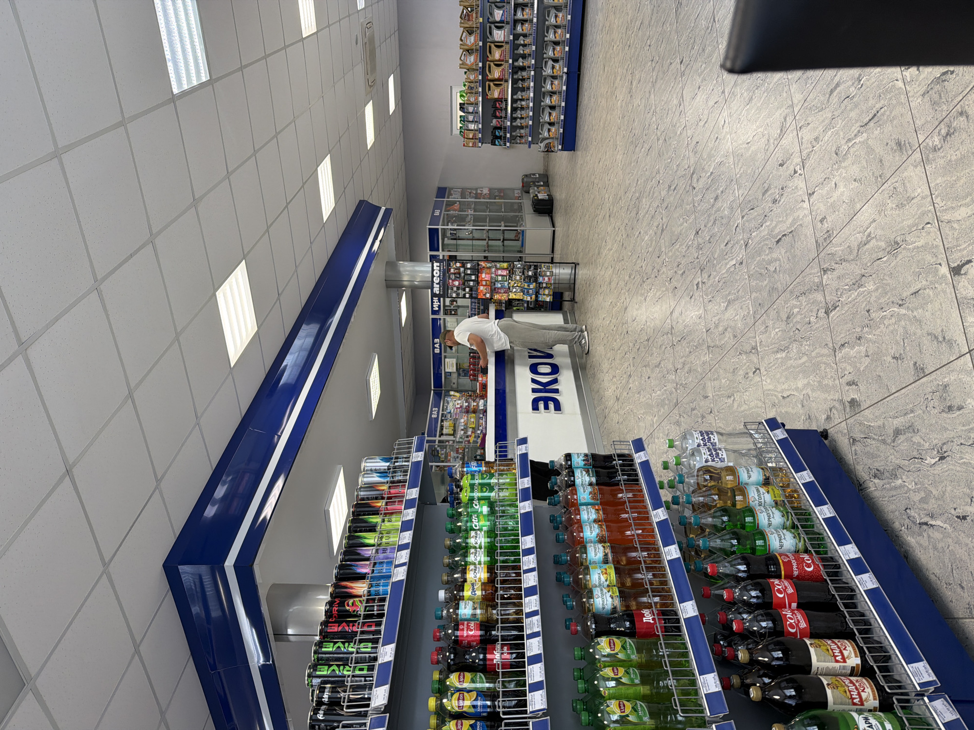 The image shows the interior of a store that sells beverages and car accessories, located in the city of Chaikovsky, Russia. A man in a white t-shirt and grey pants is standing at a counter, facing away from the camera. He is looking at products on shelves. The store has tiled floors and a white ceiling with fluorescent lighting. Shelves are stocked with various drinks, including soda bottles and energy drink cans. In the background, there are displays of car products such as oil and air fresheners. A large blue and white sign with the word "ЭКО" is visible on the counter. The overall environment appears to be a retail space.