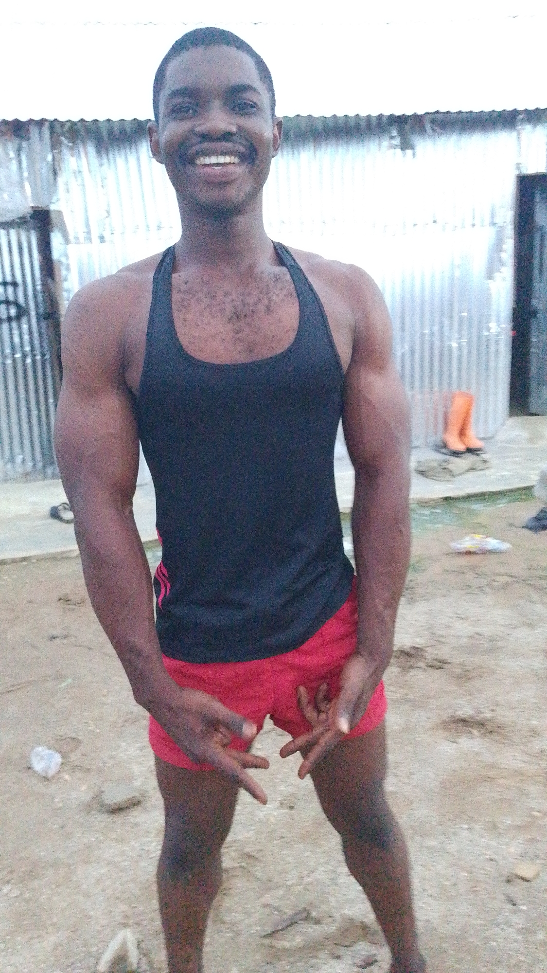A young Black male, appearing to be in his early twenties, stands outdoors in what appears to be a semi-urban or rural setting in Indo, Nigeria. He is wearing a black tank top and red athletic shorts. His physique is muscular, with well-defined arms and chest. He has a bright smile and his hands are held in front of him with his fingers splayed. The background consists of a corrugated metal wall, suggesting a simple structure or enclosure. The ground is composed of sand and dirt, with some scattered debris, including a discarded plastic bottle on the left side of the frame. In the distance, to the right, a pair of orange boots are visible. The lighting suggests it is daytime, possibly overcast.