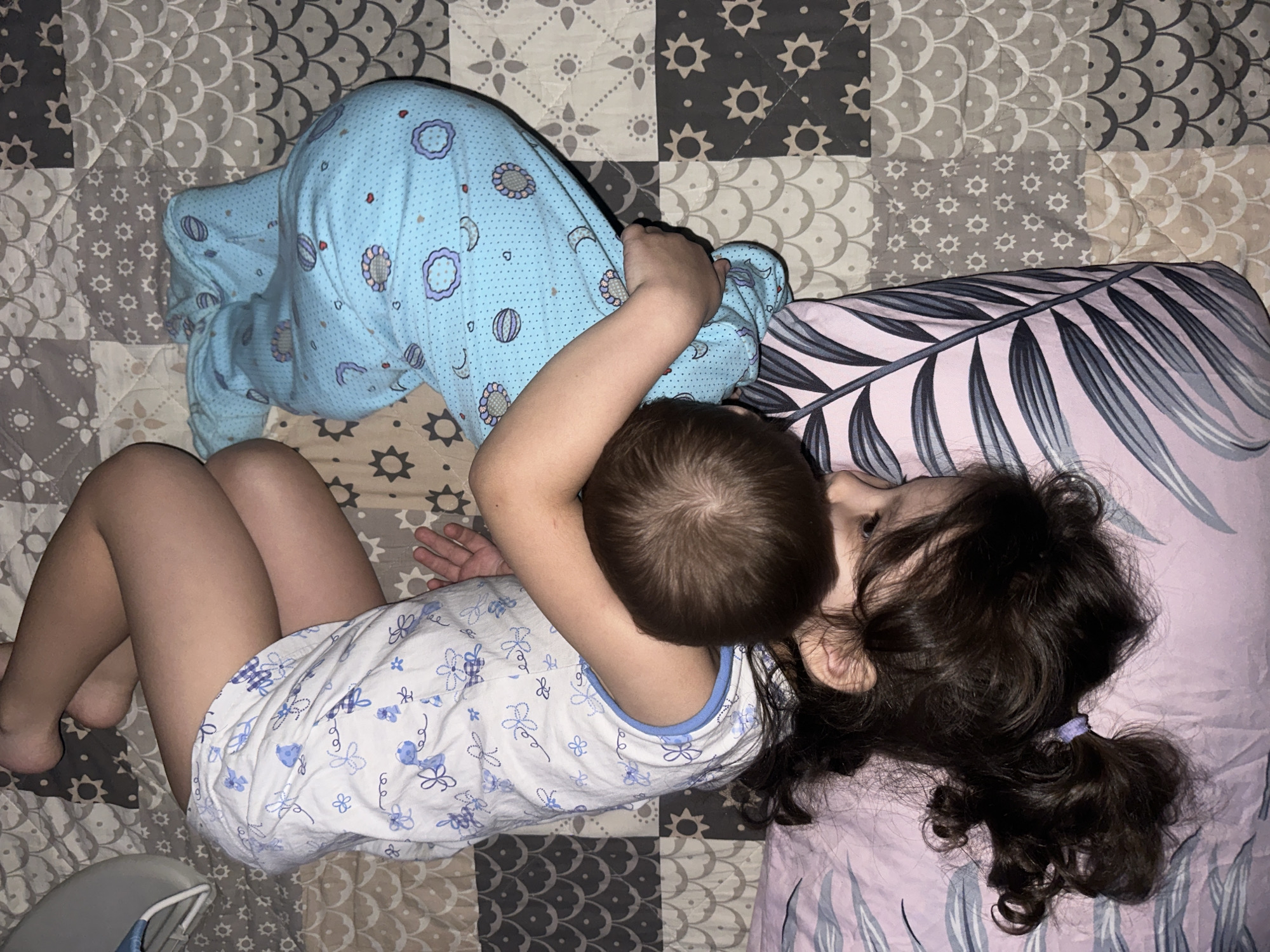 A high-angle shot shows two young children lying together on a bed with a patchwork quilt. The child on the left is facing away, wearing blue pajamas with a moon and flower design. The child on the right has curly dark hair pulled back with a purple tie, and is wearing a white short pajama set with blue flower designs. They are embracing each other. The pillow next to the second child is pink with a leaf design.