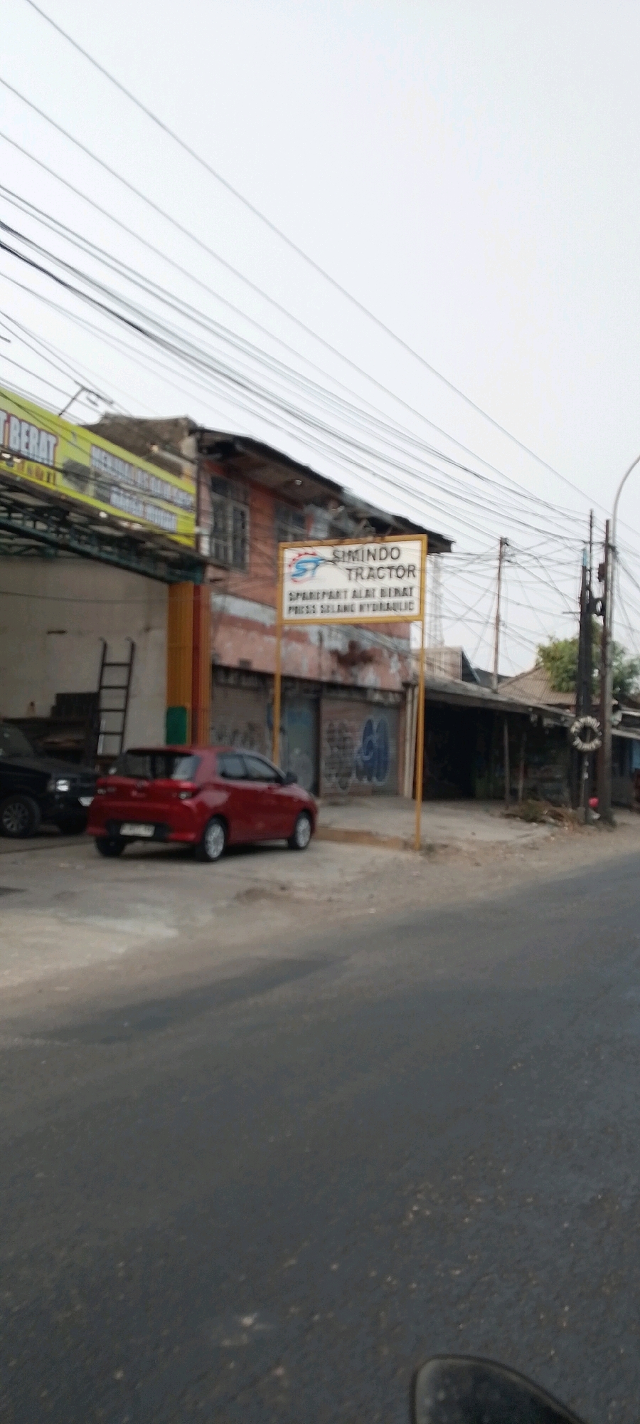 This is a medium shot, taken from street level, of a commercial building and its surroundings. The building appears to be an automotive repair shop, with a red car parked in front and a ladder leaning against the exterior. A sign above the entrance reads "SIMINDO TRACTOR" and lists "SPAREPART ALAT BERAT" and "PRESS SELANG HYDRAULIC." The building's facade is weathered, with graffiti visible on the walls. The street in front of the building is paved, and utility poles with numerous wires stretch across the frame. The sky is overcast. The scene is located in Bekasi, Indonesia, as indicated by the signage. There are no people visible in the image.