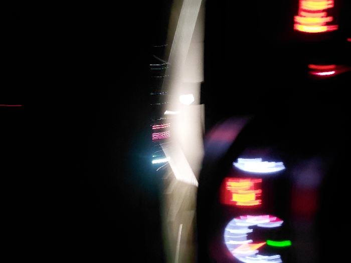 Blurred and abstracted lights dominate a black background. The right side features stacked, hazy red lights and a section with glowing numbers and a speedometer with a green indicator. A bright, vertical white streak extends from the top center, blurring into a column of light with embedded, colorful streaks. The overall image is dark and difficult to interpret.