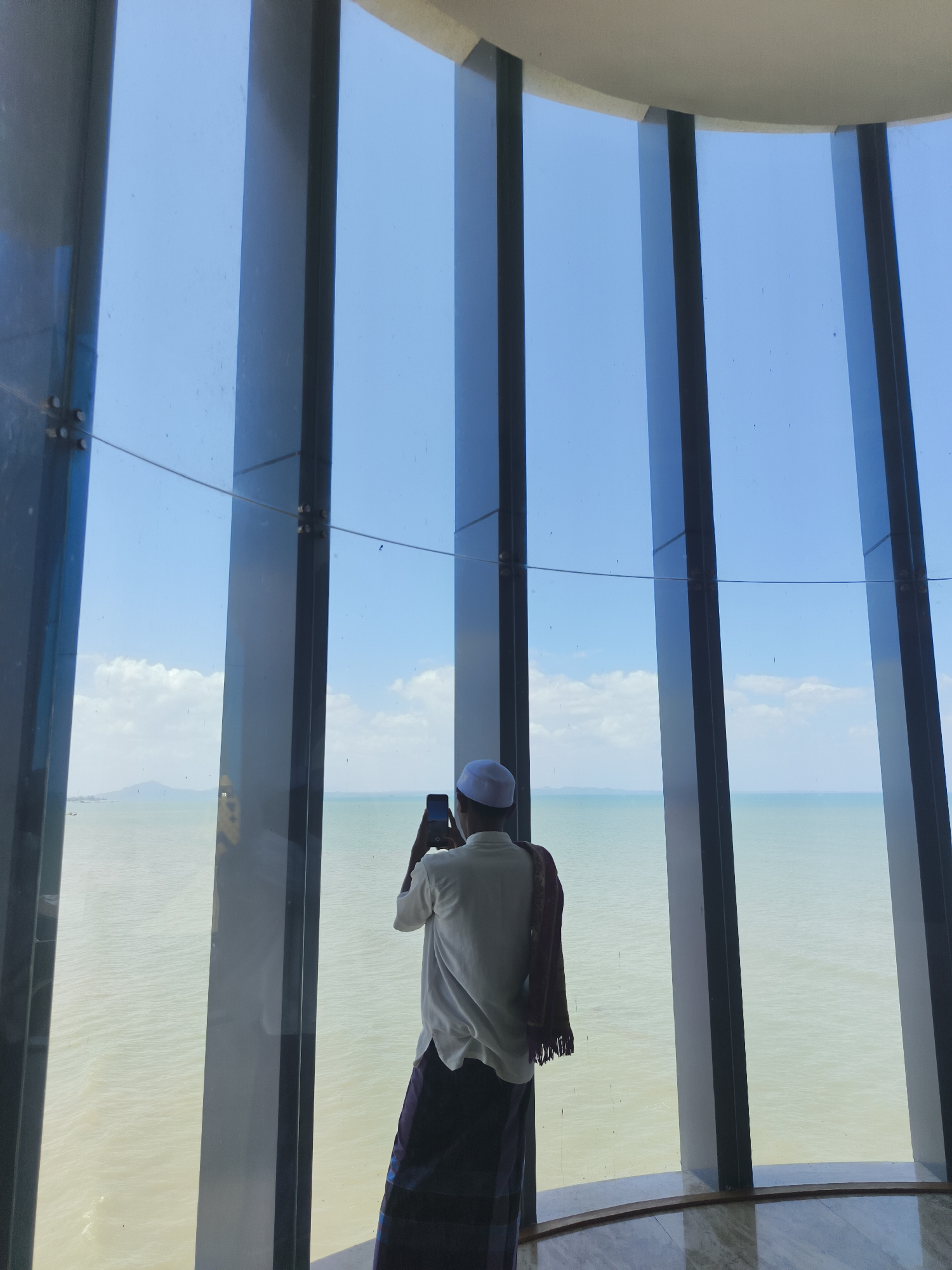 A man wearing a white kufi, white long-sleeved shirt, and a patterned sarong stands with his back to the camera, holding a smartphone up to take a photo. A maroon and white fringed cloth is draped over his shoulders. He is positioned indoors, near a large, curved window that offers a panoramic view of a calm, light-brown sea under a bright blue sky with scattered white clouds. The window is framed by tall, dark gray vertical pillars, creating a series of vertical panes. The floor appears to be made of polished marble or stone. The scene likely depicts a view from a building in the city of Beringin, Indonesia.
