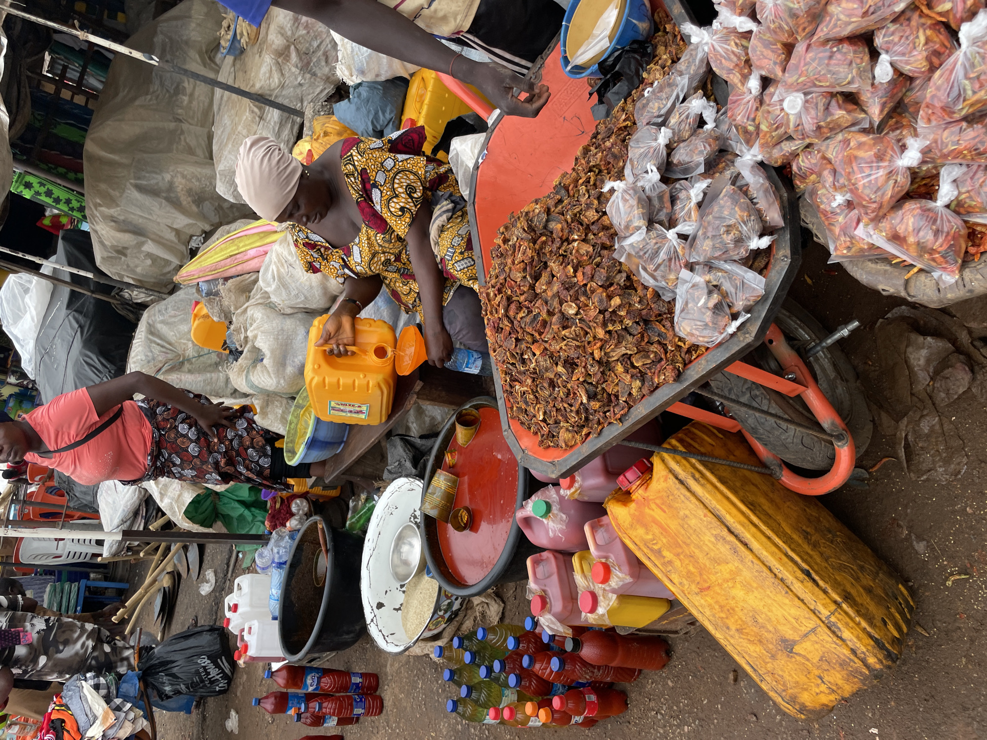The image depicts a bustling outdoor market scene, likely in Lokoja, Nigeria, characterized by a variety of food items and vendors. In the foreground, a large pile of dried chili peppers, some in plastic bags and others loose, is prominent. To the left, an array of bottled beverages, including what appears to be cooking oil and other liquids, are neatly arranged. Several large plastic containers, some yellow and others pink, hold liquids, suggesting they are for sale or transport. A woman in a vibrant, patterned dress and headscarf is seated, tending to a large orange tub filled with dried red ingredients, possibly more chili peppers or a similar commodity. She is also seen pouring a liquid from a yellow jerrycan into a smaller funnel. In the background, other market stalls are visible, with more goods covered by tarps and some shoppers and vendors moving around. The ground is dirt and appears wet in places, indicating an outdoor, possibly unpaved, market setting. The overall impression is one of a busy commercial hub where daily goods are traded.