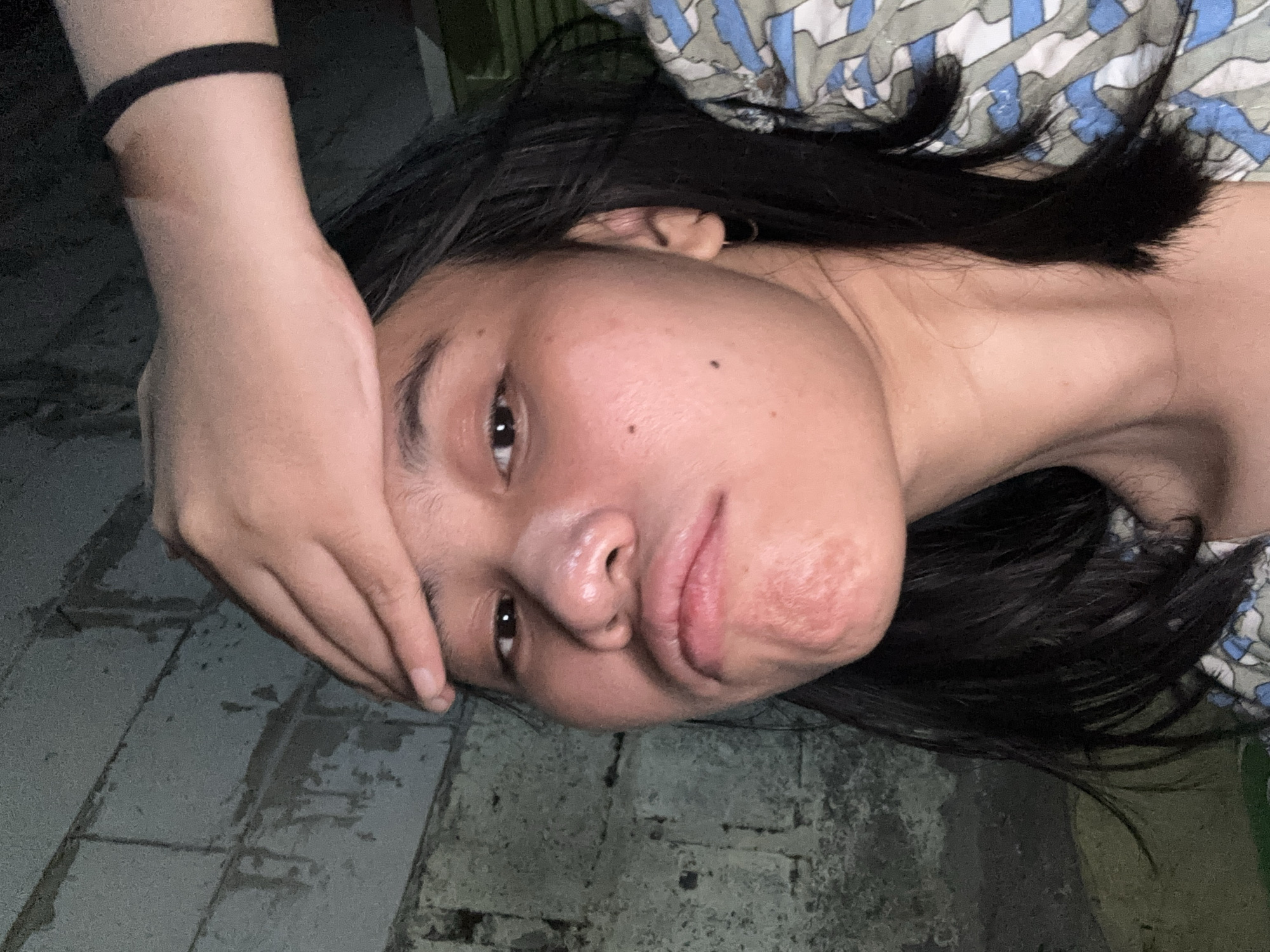 A young woman of Asian descent, with dark hair and brown eyes, is pictured indoors. She is looking at the camera with a pouty expression and her right hand is placed on her forehead. She is wearing a patterned top. The background consists of a brick wall and some green material. The lighting is dim, suggesting it is night or indoors with low light. This scene is located in the city of East Jakarta, Indonesia.
