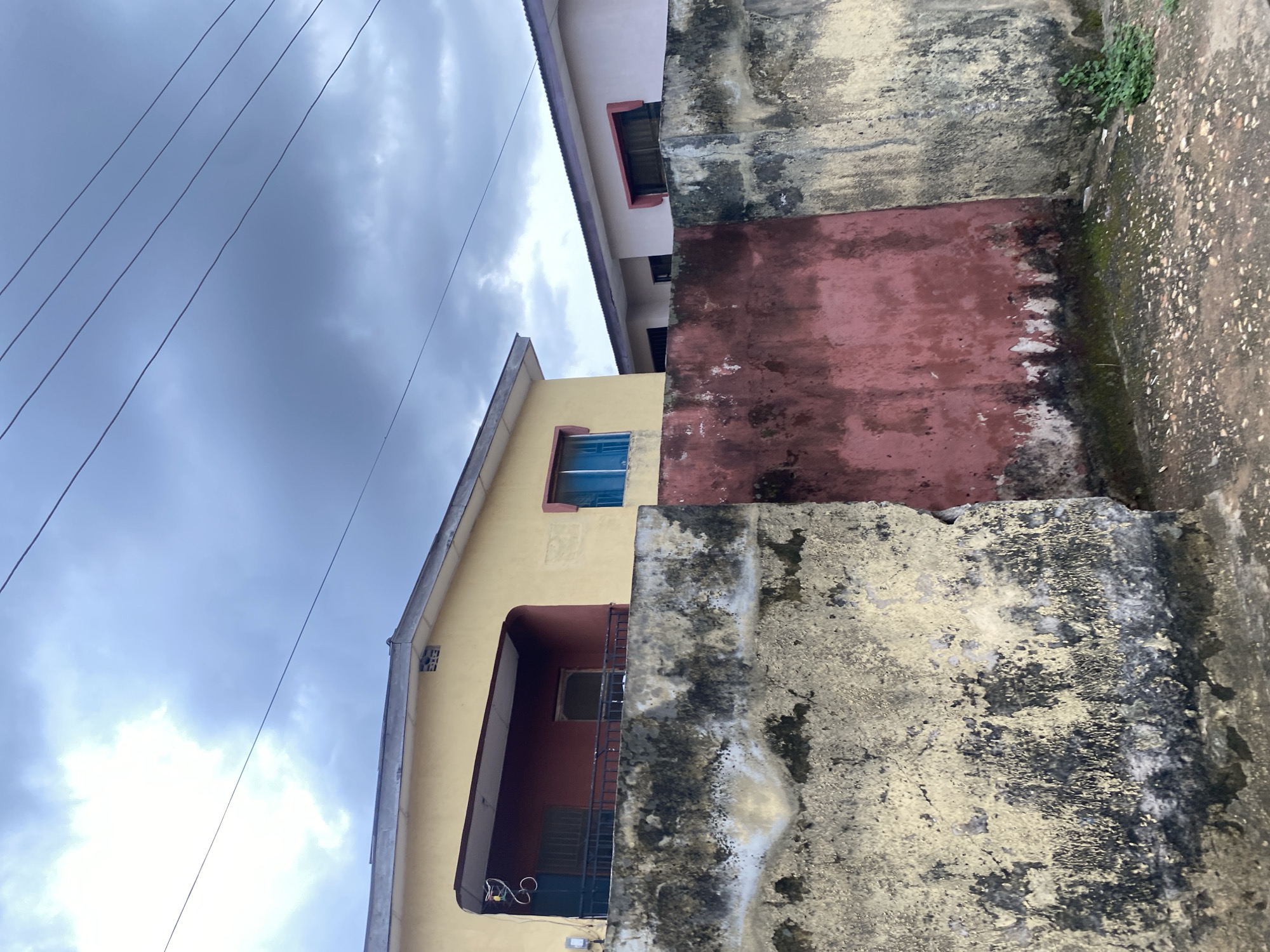 The image shows a view of buildings and walls in Lagos, Nigeria. The sky is overcast. Electrical wires are visible against the sky. A yellow-painted building with blue window frames is on the left, and a pinkish building is above it. A concrete wall, heavily stained and discolored with black and yellow, runs along the right side of the frame. Patches of moss and small plants are growing on the wall. The ground appears to be dirt or pavement. No people or actions are immediately visible.