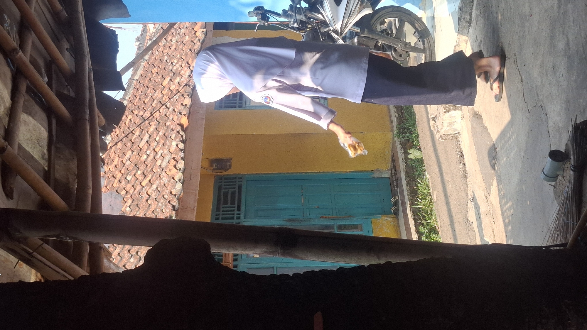 In Rahong, Indonesia, a person wearing a white hijab and a light purple uniform is standing outdoors, holding a clear plastic bag containing food items. The person is facing away from the camera, with their right hand extended, holding the bag. They are wearing dark blue pants and brown flip-flops. Behind the person is a yellow house with a turquoise door and a tiled roof. A portion of a white and blue motorcycle is visible to the right of the person. The ground in front of the house is paved with concrete and asphalt. A dark tree trunk is in the foreground on the left, and a broom is lying on the ground in the lower left corner. The lighting suggests it is daytime, with strong shadows cast by the overhead structure.