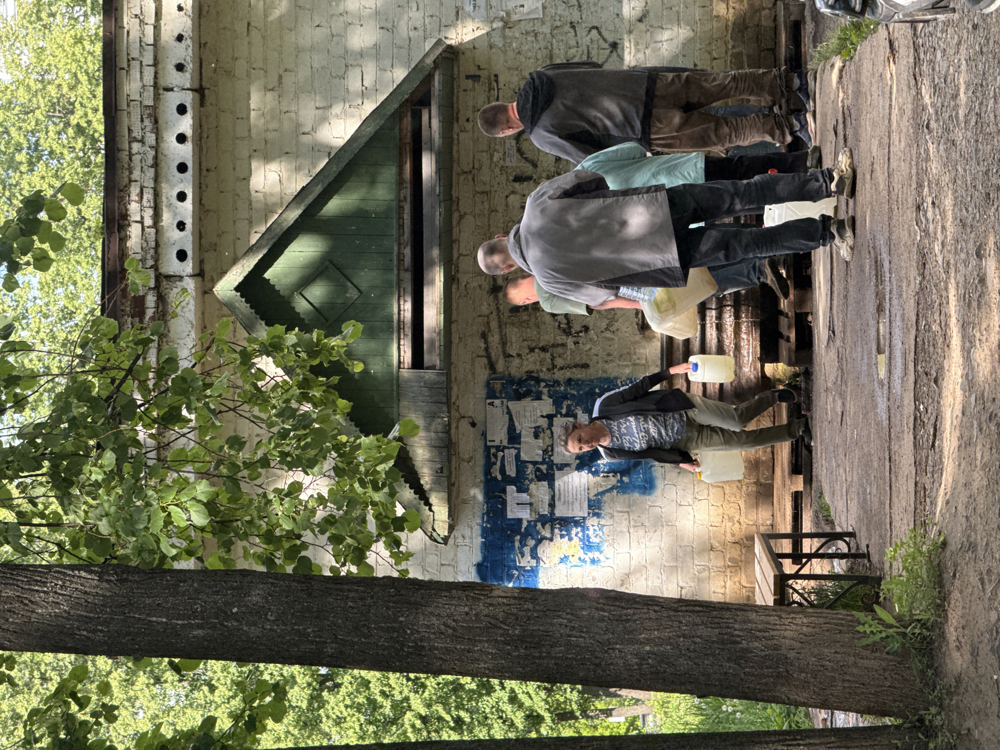This image captures an outdoor scene where several men are queuing to collect water from a public source next to an old building.
In the foreground, a large tree trunk dominates the left side of the frame, with vibrant green foliage extending above and around it. The ground, appearing to be a worn dirt or paved path, stretches across the lower portion of the image. A rustic wooden bench with dark metal supports is positioned to the right of the tree trunk.
Further back, a group of men, mostly with short hair or bald, are gathered near a water spigot or dispenser, which is not fully visible but implied by their actions. They are holding various light-colored (white or yellowish) plastic containers, suggesting they are filling them with water. One man in the immediate foreground on the right is also holding a container.
The background is dominated by an older building with a light-colored brick wall, possibly painted cream or white, showing signs of wear and age, including peeling paint and graffiti-like marks. A prominent section of the wall features a patch of blue paint covered with numerous old, torn posters and flyers. A small, dark green wooden structure with a pitched roof and a diamond-shaped detail is attached to the lower part of the building. Above this structure, a line of evenly spaced black holes is visible along a horizontal beam.
The scene is well-lit by bright daylight, casting clear shadows on the ground and the building, indicating a sunny day. The overall mood is practical and routine, centered on the everyday task of water collection. Cyrillic text is visible on some of the posters on the wall, with one partially legible word appearing to be "ПРИВІТАЙТЕ."