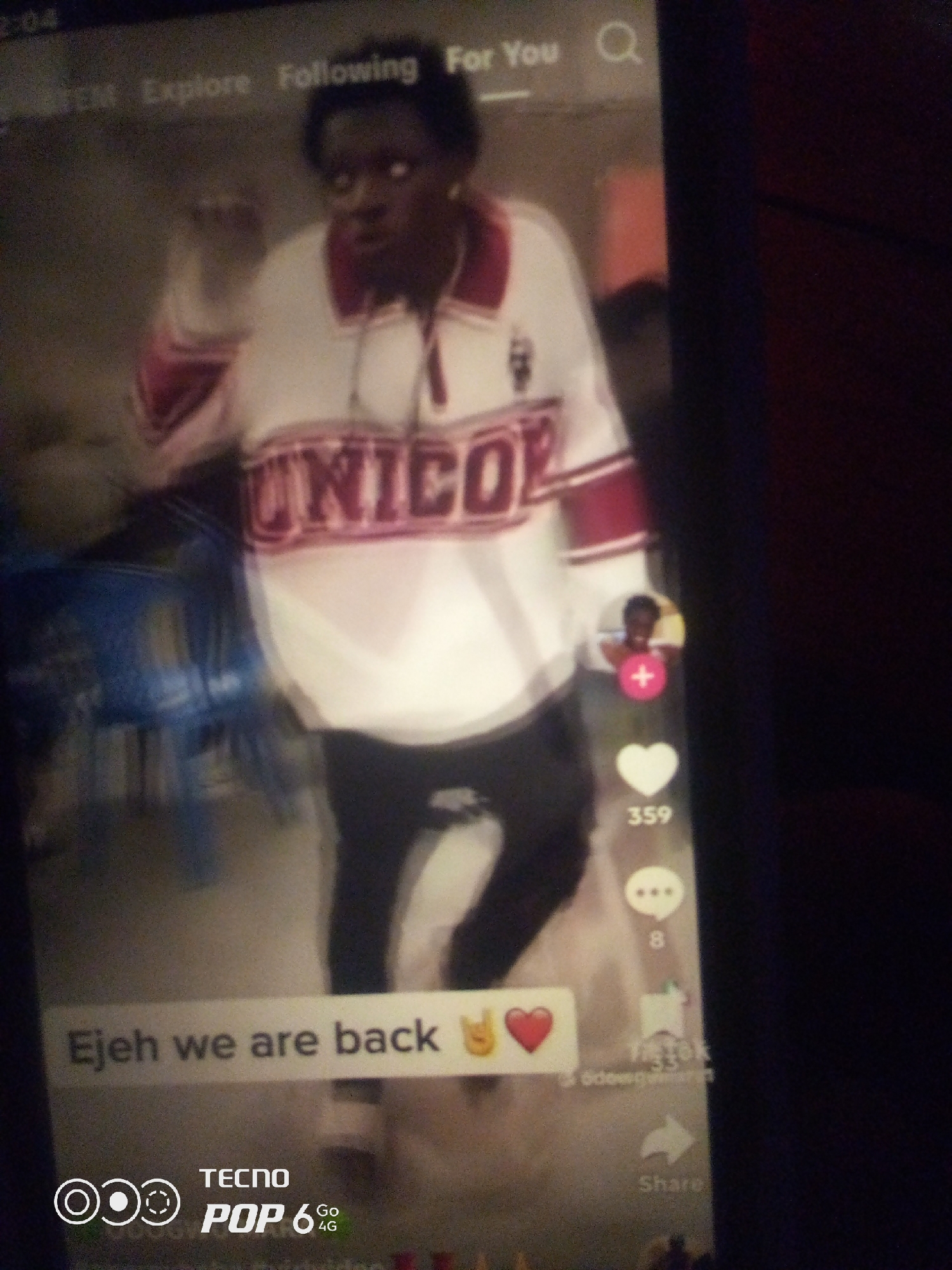 The image depicts a vertical, full-body shot of a young Black person standing and facing the camera. They are wearing a white, long-sleeved shirt with red and white stripes on the collar and sleeves, and the word "UNICORN" is prominently displayed in red letters across the chest. The background is dark and appears to be indoors, with some blurry blue chairs visible to the left. On the right side of the screen, there are typical social media icons: a profile picture with a plus sign, a heart icon with the number "359" below it, a speech bubble icon with the number "8" below it, a bookmark icon, and a share icon. Text overlaid on the image reads "Ejehh we are back" with emojis of a peace sign and a heart. At the bottom, there is a Tecno Pop 6 4G logo. The lighting is dim, and the image quality is poor, suggesting it was taken in low light conditions or is a low-resolution photograph. There is no indication of the city of Howayi, Nigeria, or any specific location within it.