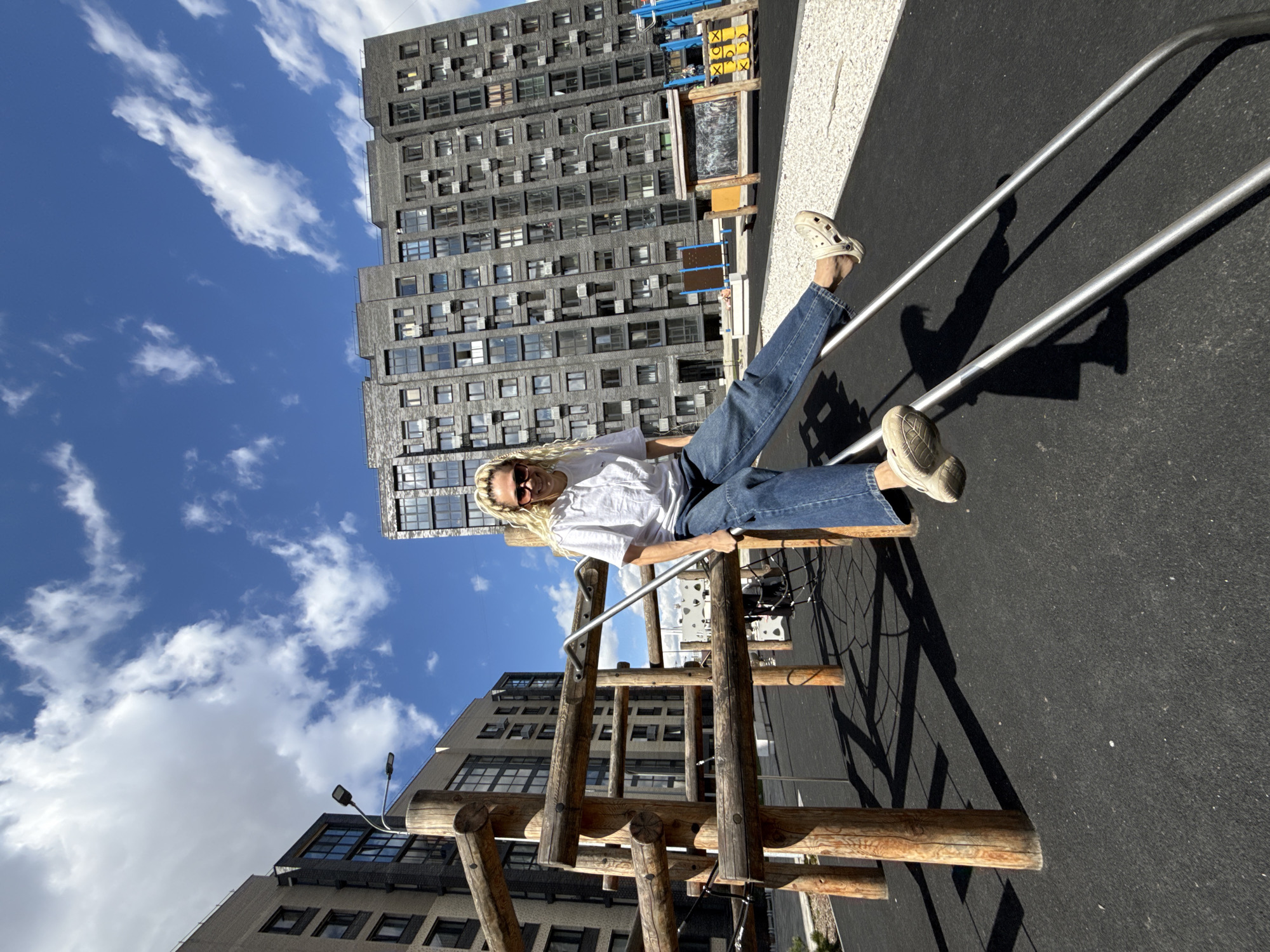 A woman wearing a white t-shirt, blue jeans, and beige footwear is sitting on a playground structure, with her legs extended. She has blonde hair and is wearing sunglasses. The playground structure is made of wooden logs and metal bars. The background includes modern buildings with multiple windows and a blue sky with scattered clouds. The location appears to be an urban area with a playground.