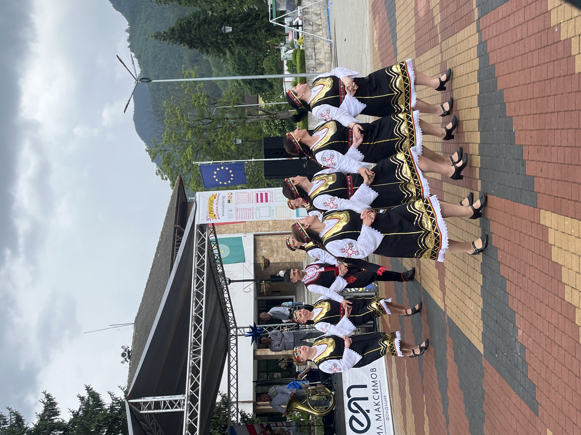 This image depicts an outdoor cultural performance in Unknown, Unknown. In the foreground, nine individuals, consisting of eight females and one male, are actively engaged in a traditional dance on a paved surface. The dancers are arranged in a diagonal line, facing the viewer's right. The female dancers are attired in black dresses with extensive gold trim, white long-sleeved blouses with embroidered cuffs, and decorative headbands featuring red and gold elements. They wear black Mary Jane-style shoes. The male dancer wears a white embroidered shirt, a black vest with red embroidery, black trousers, and shoes, with red tassels visible.
Behind the dancers, a temporary outdoor stage with a grey canopy and metal truss framework is erected. On the stage, at least two male individuals are visible; one is playing a brass instrument identified as a tuba or sousaphone, while others stand in the background. A building with a tiled roof is situated behind the stage.
Several banners and flags are displayed in the background: a partially visible blue, white, and red banner on the far left; a vertical white banner with the "em" logo and Cyrillic text including "МАКСИМОВ" and "ФОНДАЦИЯ"; a blue flag with yellow stars (European Union flag); and a green flag with an emblem. To the right of these, a tall white banner is partially visible, featuring Cyrillic text such as "ПЛАНХ", "ФАБРИКА ЗА ЩОРИ", and the web address "www.kamax.bg".
The environmental background includes dense green foliage, with both coniferous and deciduous trees, and large forested mountains rising under an overcast sky. A multi-armed street lamp is prominently positioned on the right side of the frame. Two horn-shaped loudspeakers are mounted on the roof of the building behind the stage. The ground beneath the dancers is composed of interlocking pavers in shades of reddish-brown, dark grey, and light tan, arranged in a zigzag pattern.