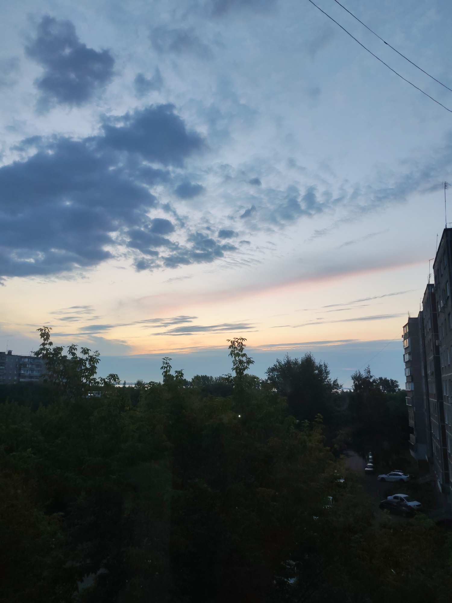 The image depicts a view from an elevated vantage point, likely an apartment window, looking out over a residential area during twilight. Dark green foliage, likely trees and bushes, dominates the foreground, obscuring much of the ground level. Beyond the vegetation, a line of apartment buildings is visible to the left and right. The building on the right is a multi-story concrete structure with visible windows and a television antenna on its roof. To the left, another apartment building is partially visible. Several cars are parked on a paved area between the buildings and the trees. The sky above is a mix of dark, heavy clouds in the upper left quadrant, transitioning to lighter, scattered clouds and a pale pink and yellow streak of light along the horizon, suggesting the sun has recently set or is about to rise. Thin power lines are visible stretching across the upper portion of the sky. The overall impression is of a typical urban evening scene in a Russian city, likely Chelyabinsk, Russia, given the architectural style of the buildings. No people are discernable in the image.