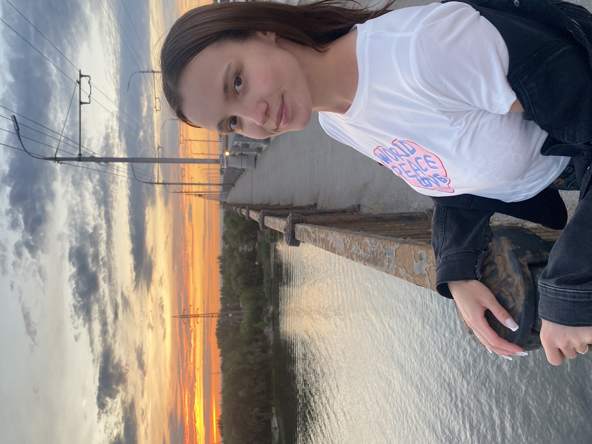 A young woman with shoulder-length dark hair stands on a bridge in Novokuybyshevsk, Russia, looking towards the camera with a slight smile. She is wearing a white t-shirt with pink and blue lettering that reads "WORLD PEACE LOVE" and a black denim jacket draped over her shoulders. Her hands rest on a weathered metal railing, showcasing white-tipped manicured nails and silver rings.
The bridge spans a calm body of water, reflecting the soft hues of a sunset. The sky is a canvas of oranges, yellows, and grays, with scattered clouds and utility poles stretching into the distance. Trees line the far bank of the water, and the bridge itself is lined with lampposts and overhead wires. The perspective suggests the photograph was taken at eye level, with the woman slightly off-center to the right. The overall atmosphere is serene and picturesque, capturing a moment at dusk.
