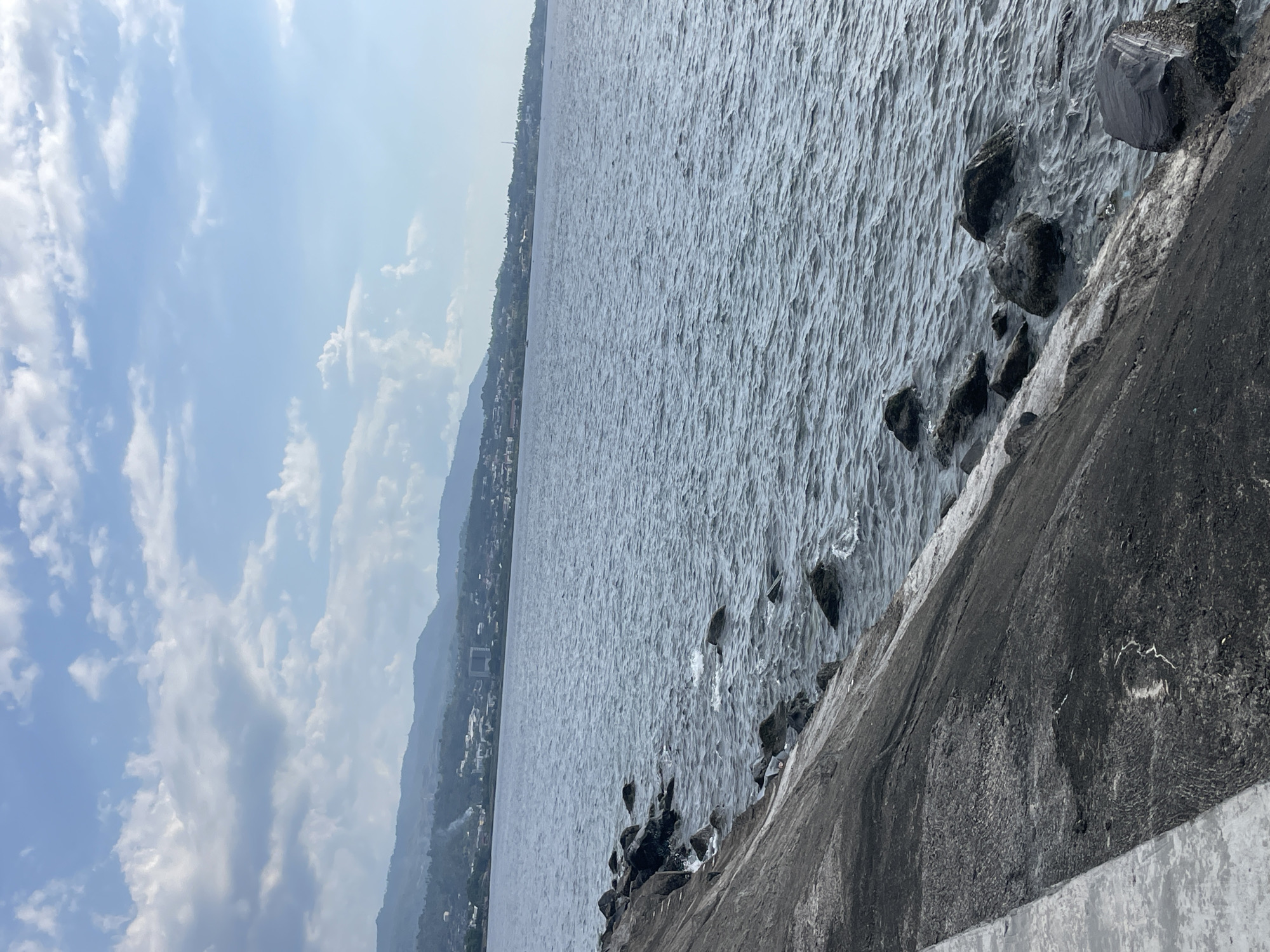The image depicts a body of water with a rocky shoreline. In the background, a city with buildings and hills can be seen. The sky is partly cloudy with a blue hue. The scene is likely a coastal area in Manado, Indonesia, given the typical landscape and climate. There are no people or animals visible in the image. The focus is on the natural elements of the sea and land, with the city serving as a backdrop.