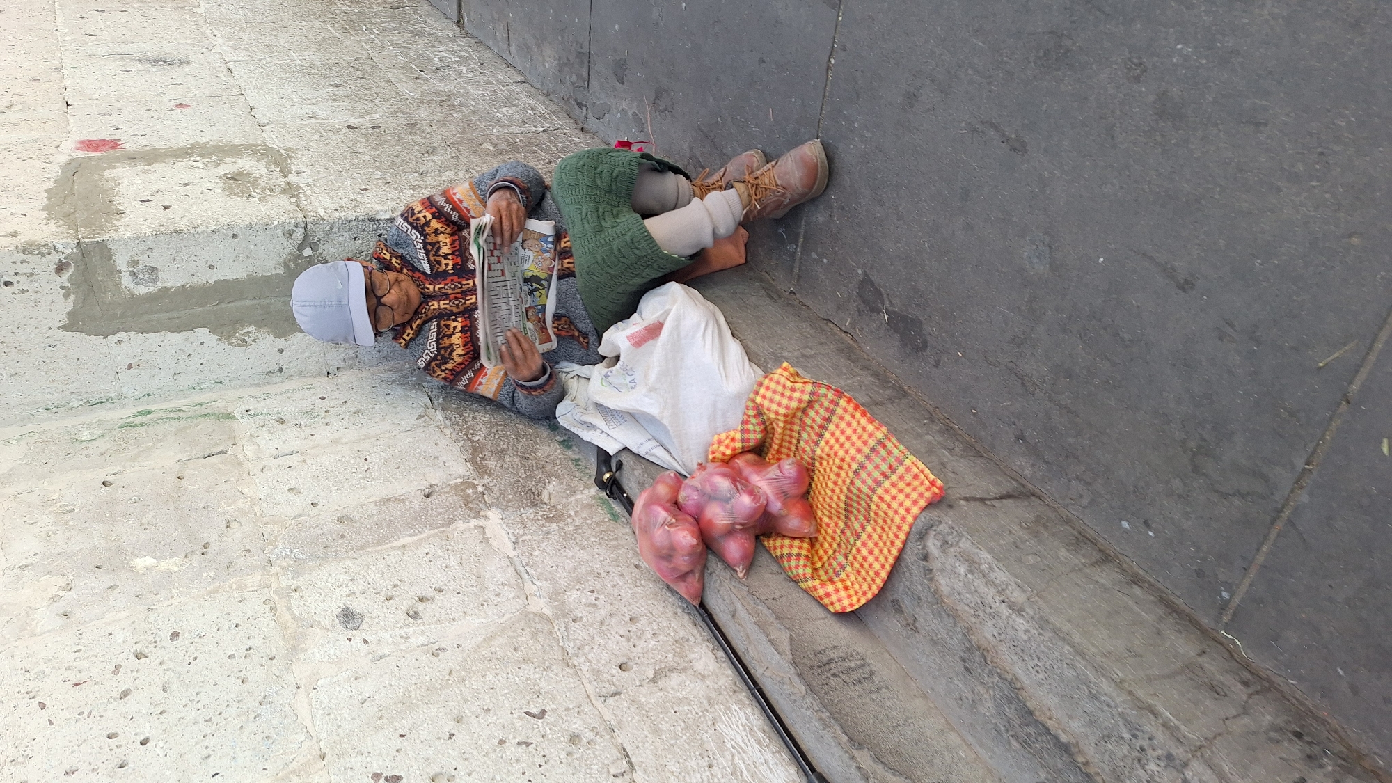 A person is lying on the ground next to a concrete wall in an urban environment. The person is wearing a gray cap, eyeglasses, and a patterned sweater, and they are reading a newspaper. A bag of onions, a white bag, and a checkered cloth are placed near the person. The ground is made of stone or concrete. The person is lying partially in a recessed area or drainage channel in the ground.