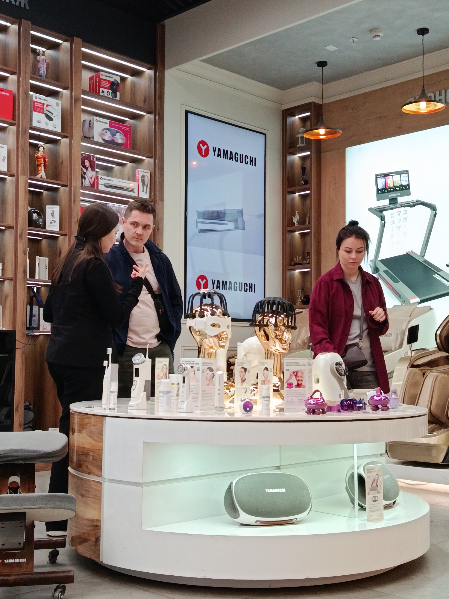 The image shows the interior of a Yamaguchi store, likely located in Moscow, Russia, based on the Cyrillic text on a nearby screen and the brand's known presence in Russia. A saleswoman in a black jacket is engaging in conversation with a man in a navy jacket. A woman in a red shirt stands on the other side of a circular display table, examining products. The table is stocked with various beauty and massage devices. In the background, there are shelves lined with Yamaguchi products and promotional screens displaying the brand's logo. A massage chair and a treadmill are visible in the background on the right side of the image. Lighting fixtures hang from the ceiling.
