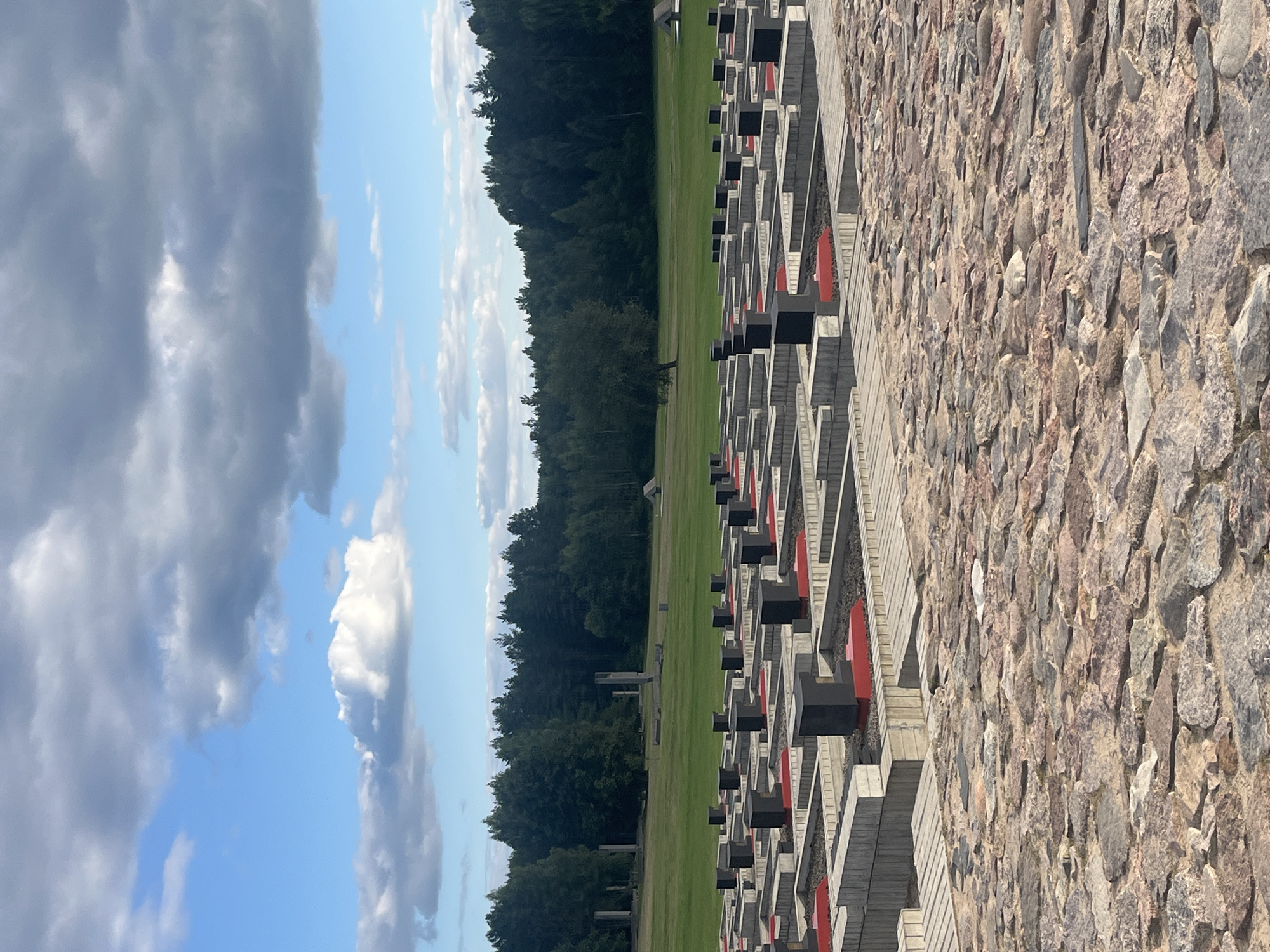 The image shows a memorial complex located in the city of Kamenski Selski Savet, Belarus. The foreground is paved with irregular, reddish-brown stones. In the mid-ground, a vast memorial area consists of numerous rectangular concrete structures arranged in a grid pattern. Each structure has raised edges and contains dark, cuboid markers, some of which are placed atop red rectangular bases. Beyond the memorial, a wide expanse of green grass stretches towards a dense line of dark green trees. In the distance, the trees are interrupted by remnants of stone structures, possibly ruins or historical monuments, and a small, triangular roofed structure. The sky above is blue with scattered white and gray clouds.