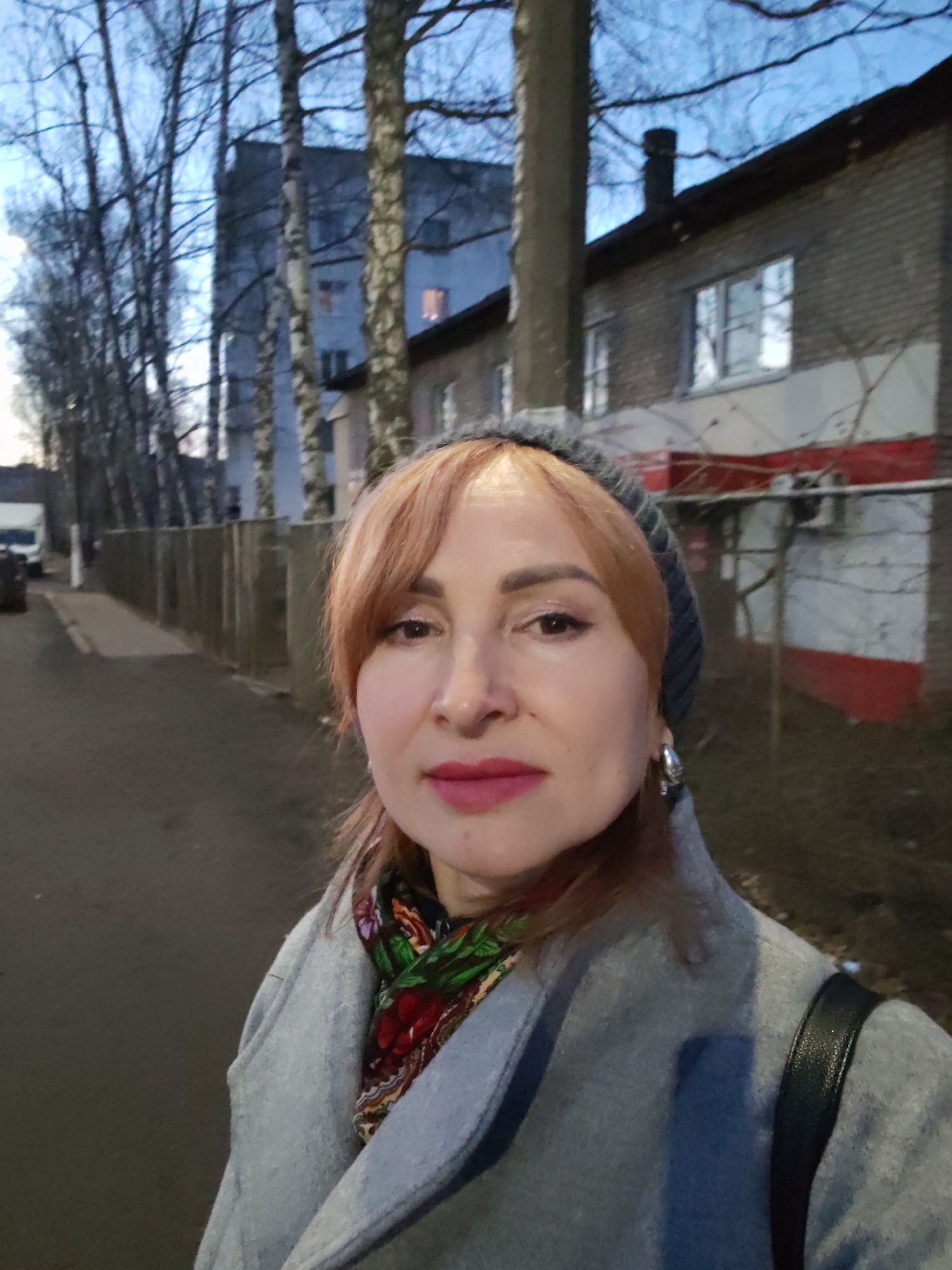 A woman is captured in a close-up outdoor portrait, likely a selfie, on a street in Chaikovsky, Russia. She has reddish-blonde hair with bangs, wears a dark grey knitted beanie, a light grey coat, and a vibrantly patterned scarf with red and green floral motifs. Her makeup includes pink lipstick and eye makeup, and she has a silver hoop earring on her visible left ear. Her expression is calm, with a slight, subtle smile.
The setting is an urban or suburban street during dusk or early evening, indicated by the soft, fading light and the blue hue of the sky. Lights are visible in the windows of some background buildings. The woman's warm attire, including a coat, hat, and scarf, suggests cool or cold weather.
In the background, to the left, a long wooden fence runs alongside the street, leading to a line of bare trees, possibly birches, and a multi-story residential building in the distance. A white van and another vehicle are parked further down the street. To the woman's right, a brick building with white window frames features sections painted white and red, and what appears to be an outdoor unit on its wall. The architecture and trees are consistent with the provided location context. No visible text is present in the image.