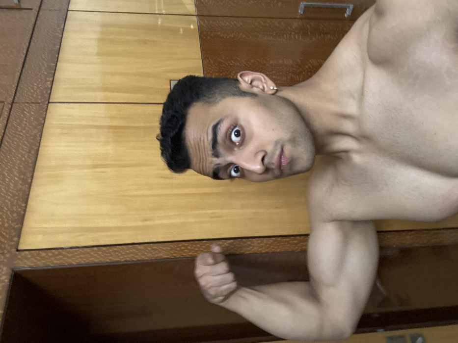 A man with short black hair and a light skin tone poses shirtless, flexing his bicep. He wears a small gold hoop earring in his left ear. His eyes are wide and he is looking at the camera with a slightly surprised expression. The background consists of light brown wooden furniture with a decorative pattern along the edges. The image is oriented horizontally.