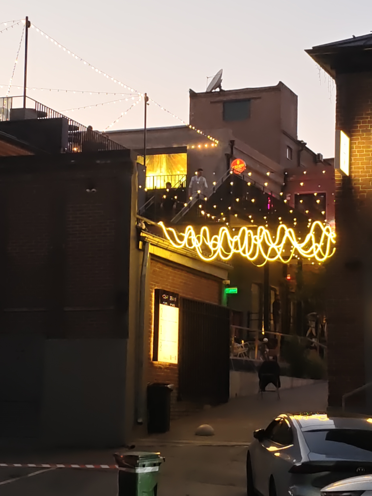 The image depicts an outdoor scene in the city of Rostov-on-Don, Russia, likely at dusk or early evening. In the foreground, a white car is parked. To the left, a green trash bin is visible, with red and white striped tape marking a boundary. The background features a narrow alleyway between brick buildings. String lights, some in a wavy pattern, are illuminated and draped across the alley, adding a festive atmosphere. People are visible on elevated walkways and stairs, suggesting the presence of restaurants or bars. One person is seated on a chair further down the alley. The architecture includes brick and concrete structures, and there is a satellite dish on one of the rooftops. The sky is a pale orange, indicating the end of daylight.