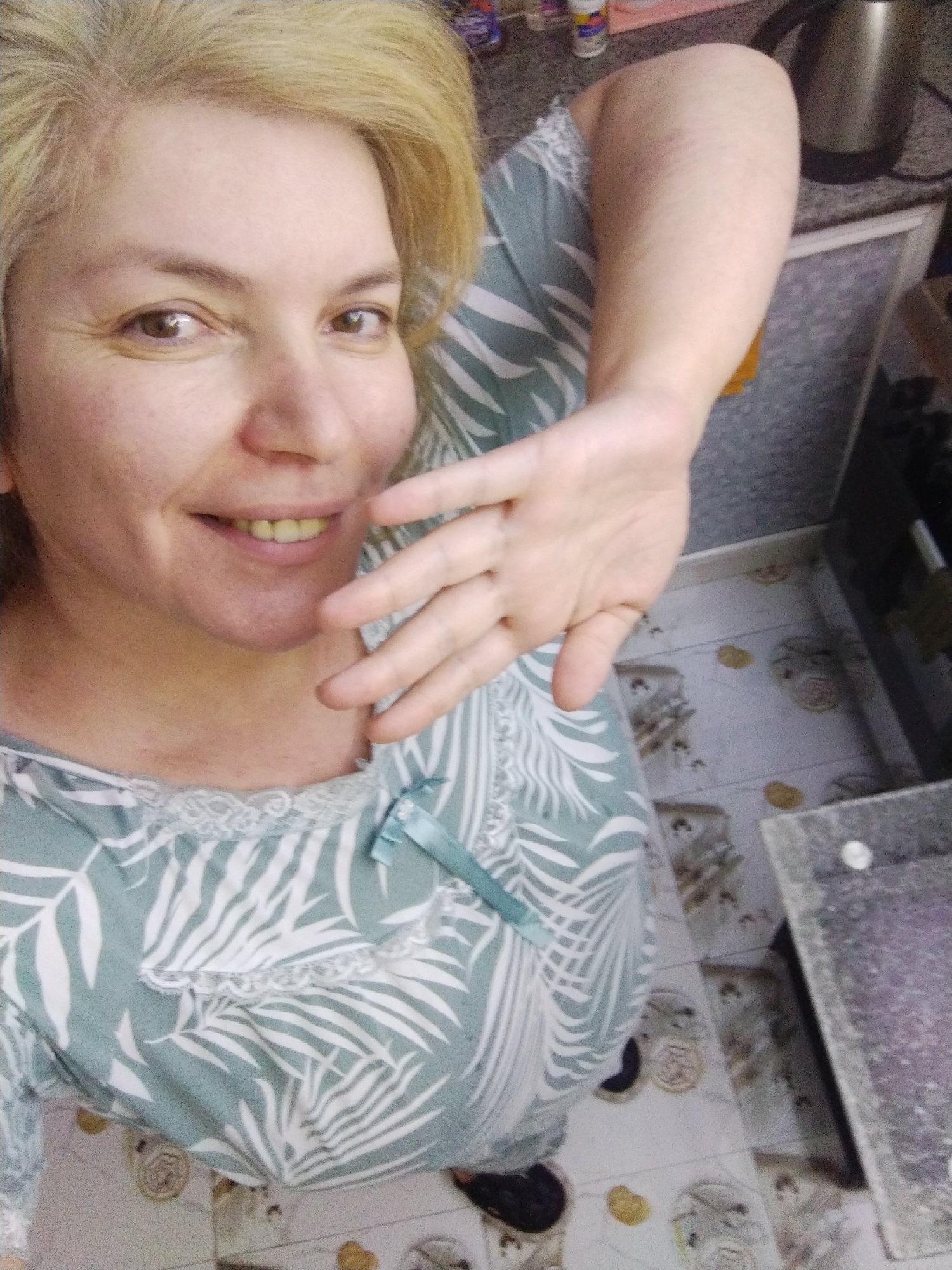 The image shows a woman with blond hair smiling and holding up her hand in a waving gesture. She is wearing a light green and white patterned top with a lace trim and a small blue bow. She appears to be standing in a kitchen, as evidenced by the tiled floor, cabinets, and a kettle on the counter in the background. The floor tiles have a pattern with what appear to be floral or medallion designs. The specific location in Al Mas'oudi, United Arab Emirates is not discernible from the image.