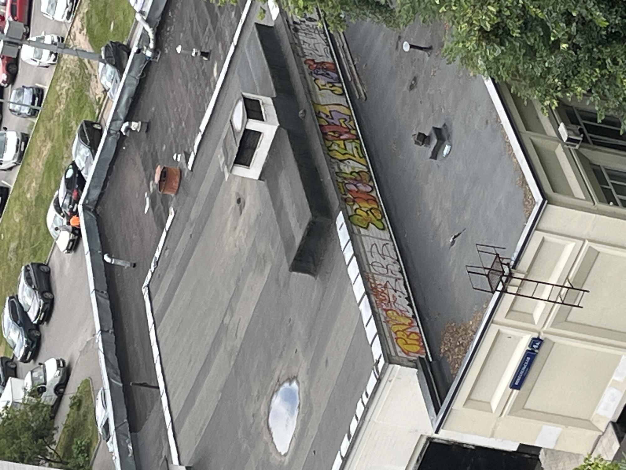 The image shows a rooftop scene in Moscow, Russia. The rooftop is flat and covered with tar. There are several objects on the rooftop, including a small white structure, a rusty barrel, ventilation pipes, and a ladder. Graffiti covers the side of the building that runs along the rooftop. In the background, there are cars parked on a street and buildings. The sky is overcast.