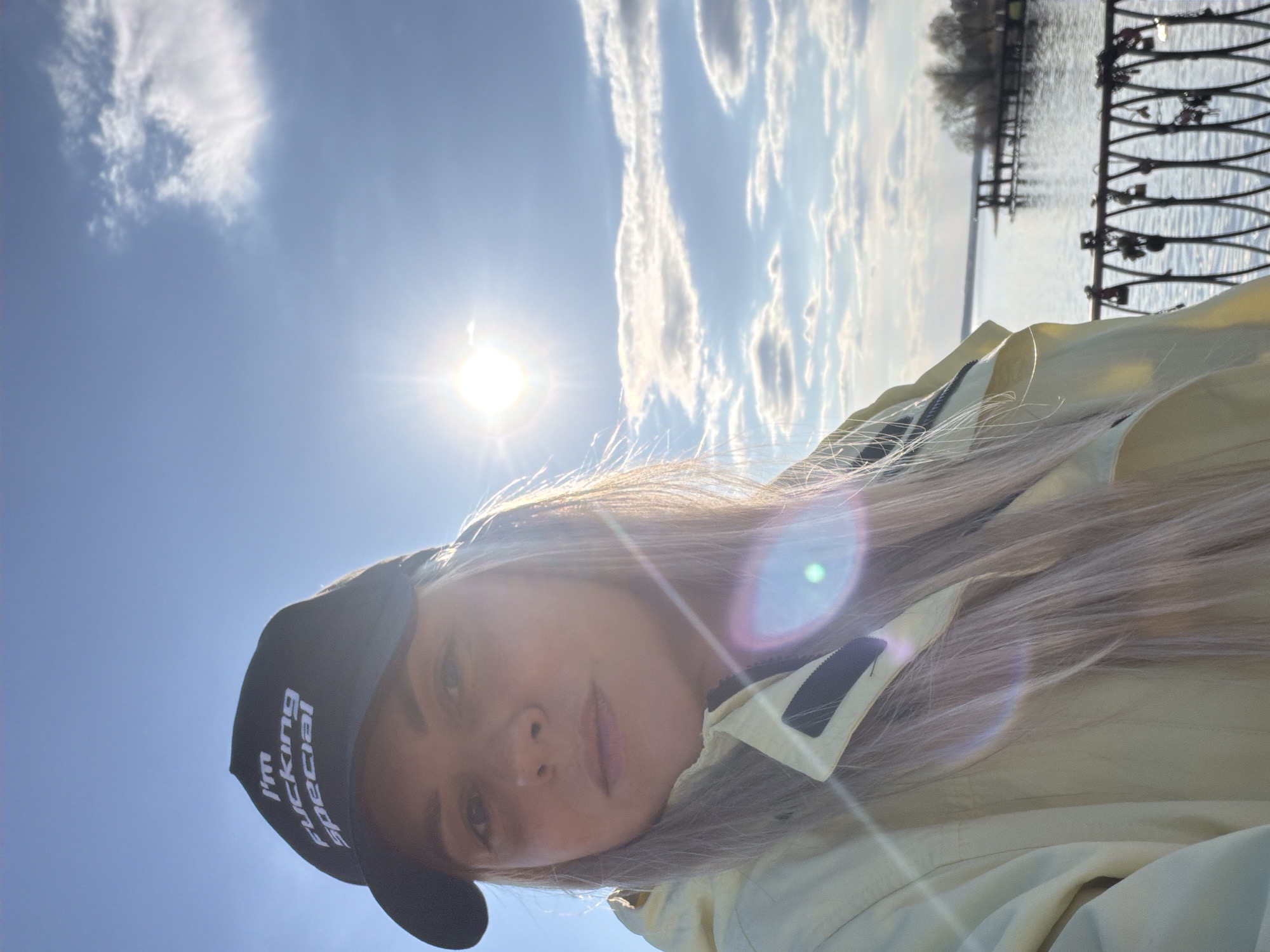 A young woman is taking a selfie. She is wearing a black cap with white lettering, a light-colored jacket, and has long, light-colored hair. The text on her hat reads "I'm fucking special." She is outdoors with a body of water and a decorative wrought-iron fence visible in the background, with trees in the far distance. The sky is blue with clouds and the sun shining brightly. The location is Votkinsk, Russia, based on the provided context.