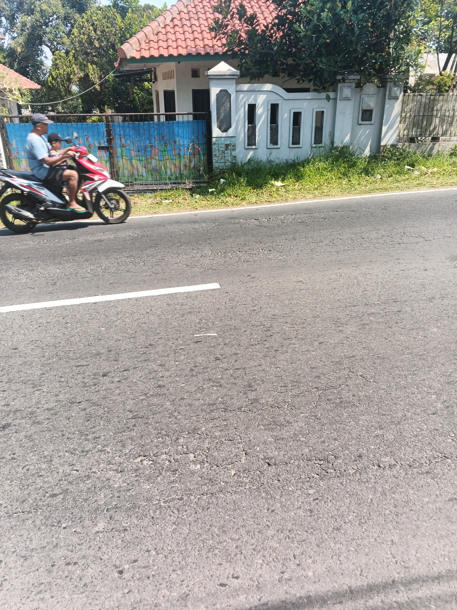 In Bandungan, Indonesia, a man and a child ride a red and white motorcycle on a paved road. The man, wearing a blue t-shirt and a cap, is driving, while the child, in a striped shirt, sits behind him. They are passing by a property with a blue gate adorned with colorful drawings and a white fence with rectangular openings. Behind the fence, a house with a red tiled roof and green trees are visible. The road has a white line marking and appears to be in moderate condition with some cracks and wear. The scene is bathed in sunlight, indicating a daytime setting.