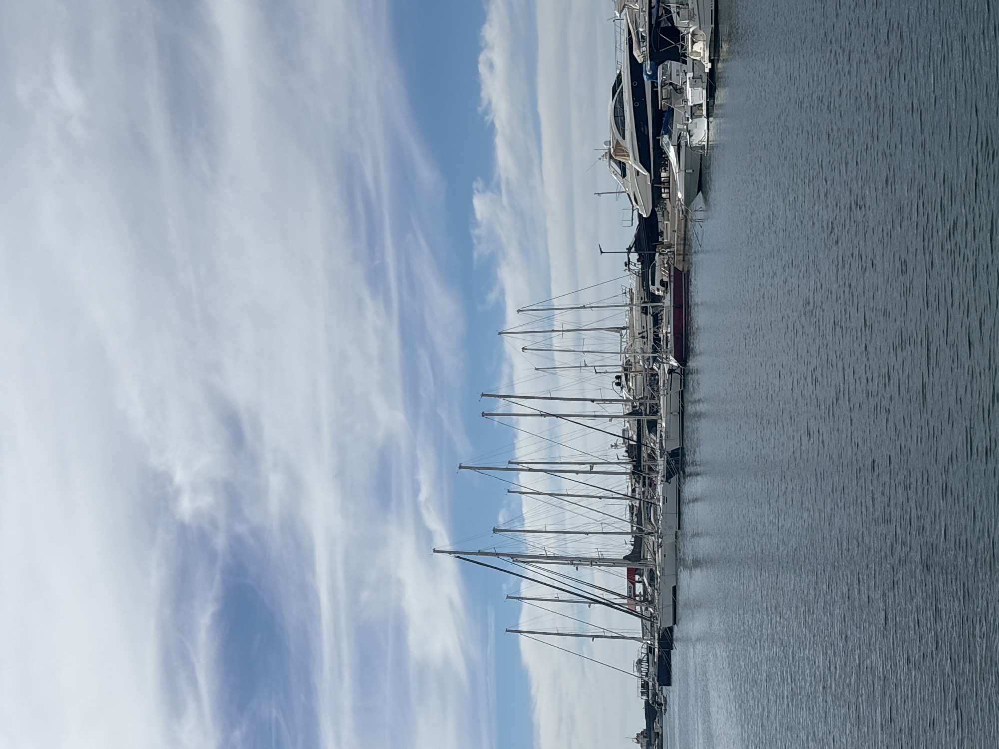 The image captures a row of sailboats and yachts moored in the harbor of Bar, Montenegro, under a bright, blue sky streaked with cirrus clouds. The masts of the sailboats are prominent, reaching upwards, while larger yachts are docked nearby. The water surface has slight ripples. The image is shot from a low angle.