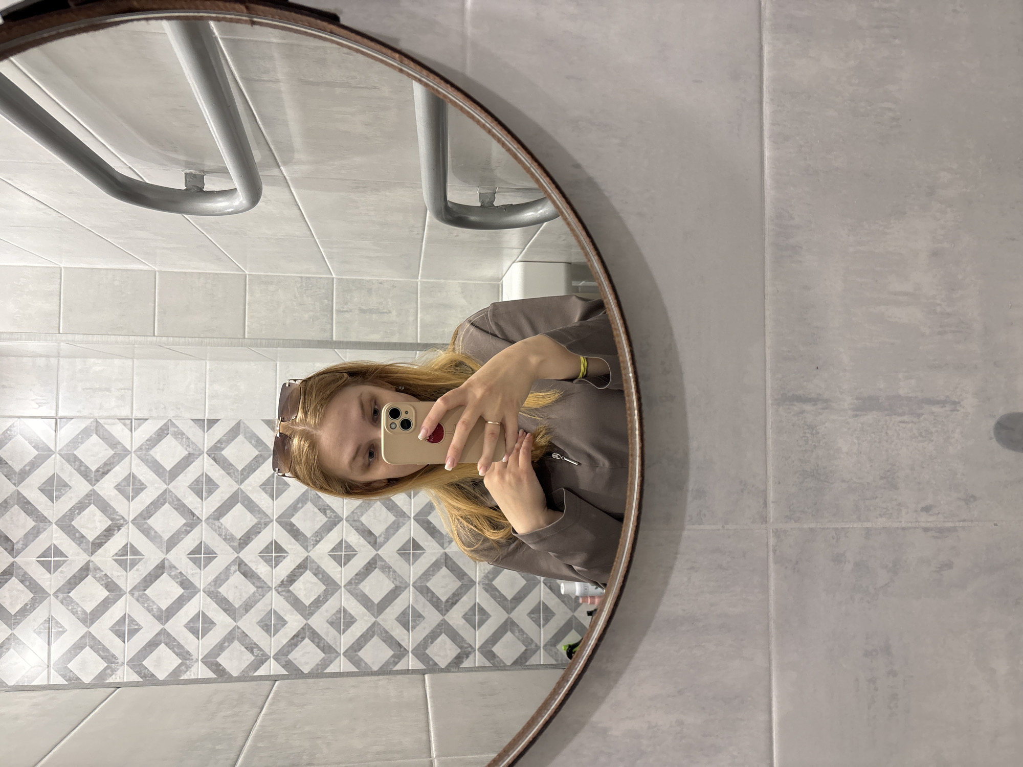 A woman takes a selfie in a round mirror. The reflection shows a bathroom with grey and white diamond pattern tiles on the wall behind her. Above this wall are white, rectangular tiles, and a chrome towel rack. To the right of the mirror's reflection is a wall with long, light grey tiles. The location may be in Podosenikha, Russia.