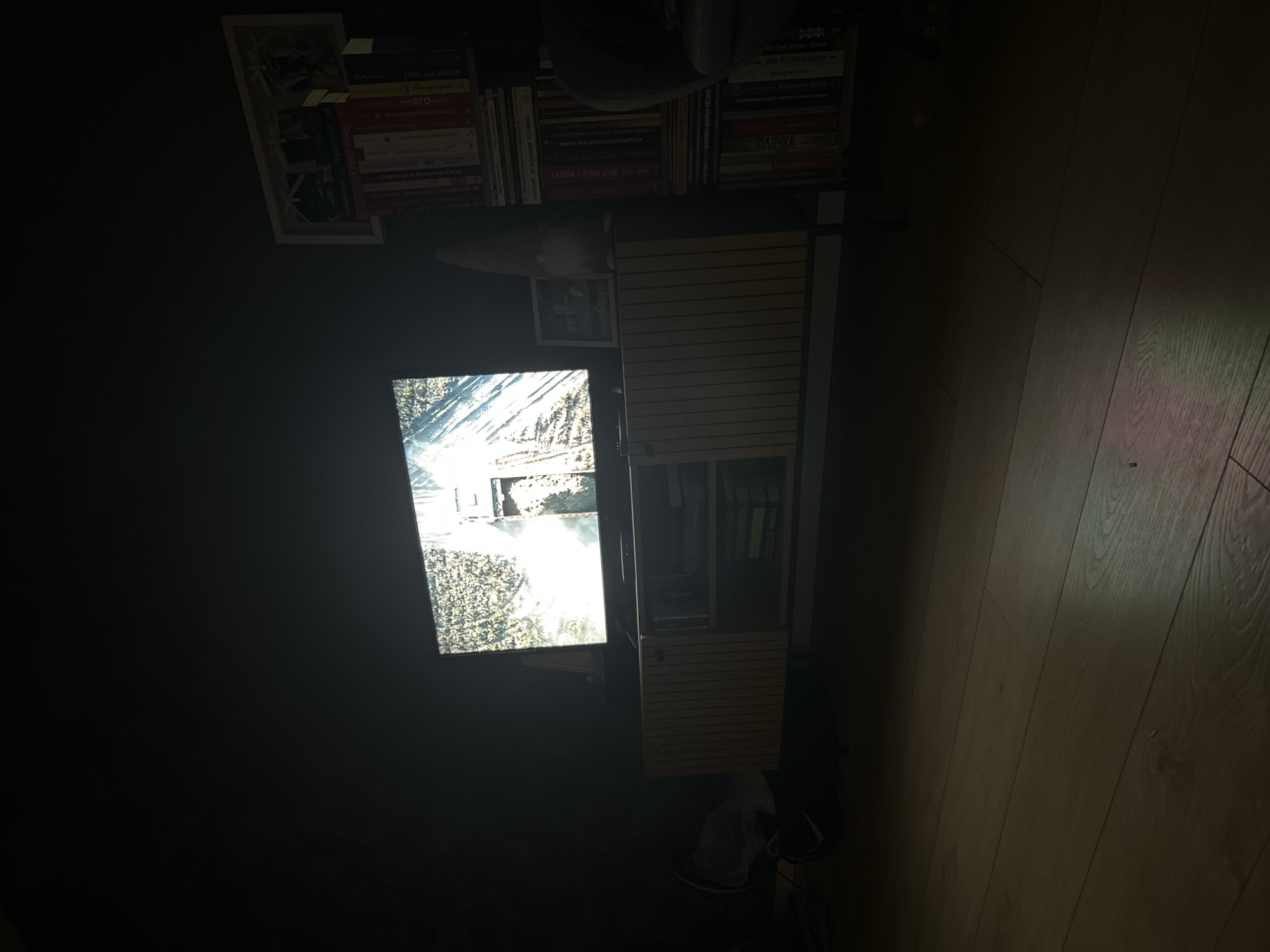 The image shows a darkened room with a television displaying a blurred image. A wooden cabinet stands to the right of the TV. Above the cabinet is a shelf with books. On the floor to the right, there are wooden planks. The lighting is dim, with a streak of light illuminating a section of the floor. The image provides no specific information to confirm the location being Plovdiv, Bulgaria.