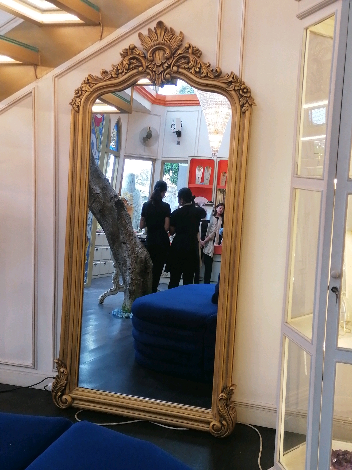 The image shows a full-length ornate golden mirror reflecting a store interior. Two women, dressed in black, are visible in the reflection, one with her back to the camera, and another facing slightly away. A third woman, wearing a beige cardigan, stands to the right of them. In the background, shelves display jewelry. A large tree trunk stands behind the mirror, leaning into the frame, and a blue cushioned seating area is visible in the foreground, partly obscured by the mirror. The scene appears to be inside a boutique in Ubud District, Indonesia, suggested by the style of the decor and the presence of what looks like a traditional Balinese-style display cabinet on the right.