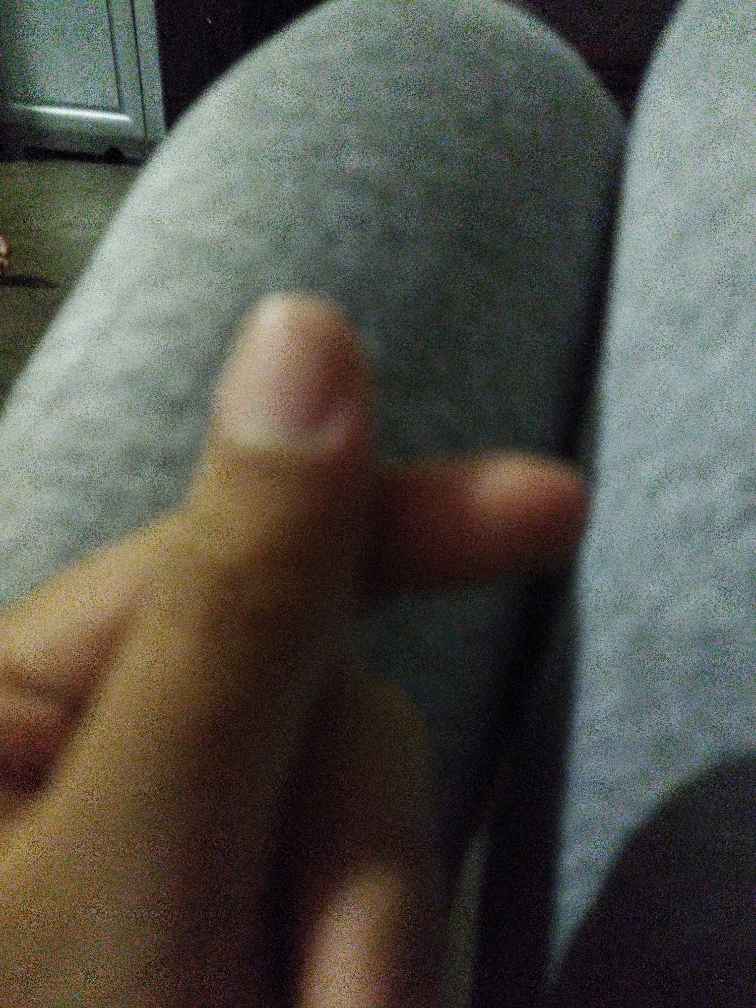 An extremely grainy, out-of-focus, close-up shot depicts a person's hand forming a Korean heart symbol. The background is dominated by a soft, gray, textured surface, likely clothing. To the left, a blurred, metallic object, possibly furniture, is visible. The scene appears to be indoors, in a dimly lit environment. No specific location in West Jakarta, Indonesia, can be identified from the provided image.