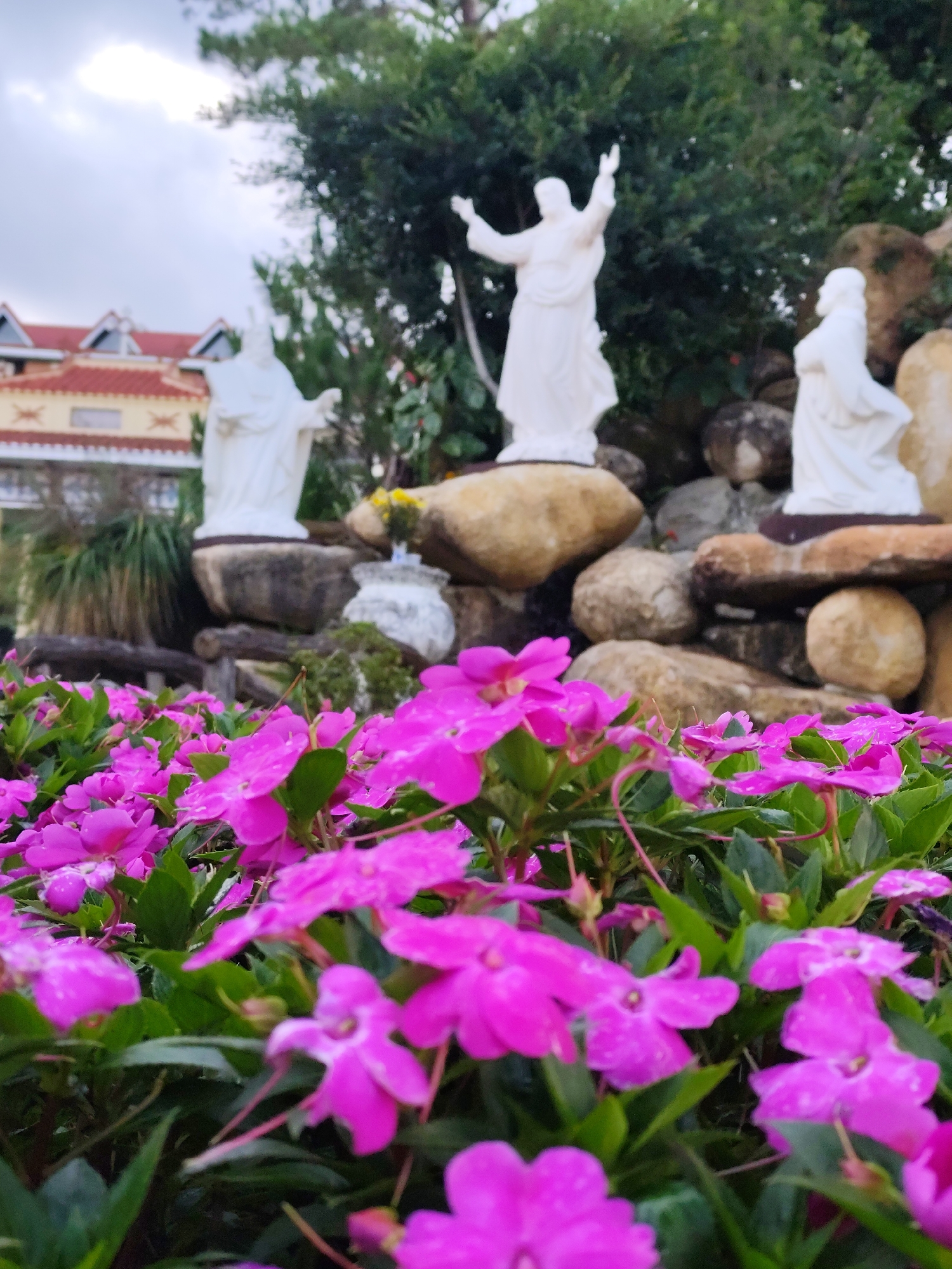 This is a medium shot taken in Unknown, Unknown, featuring a flower bed filled with vibrant pink flowers in the foreground. Behind the flowers, a small rock garden is visible, adorned with three white statues of people. A building can be seen in the background.