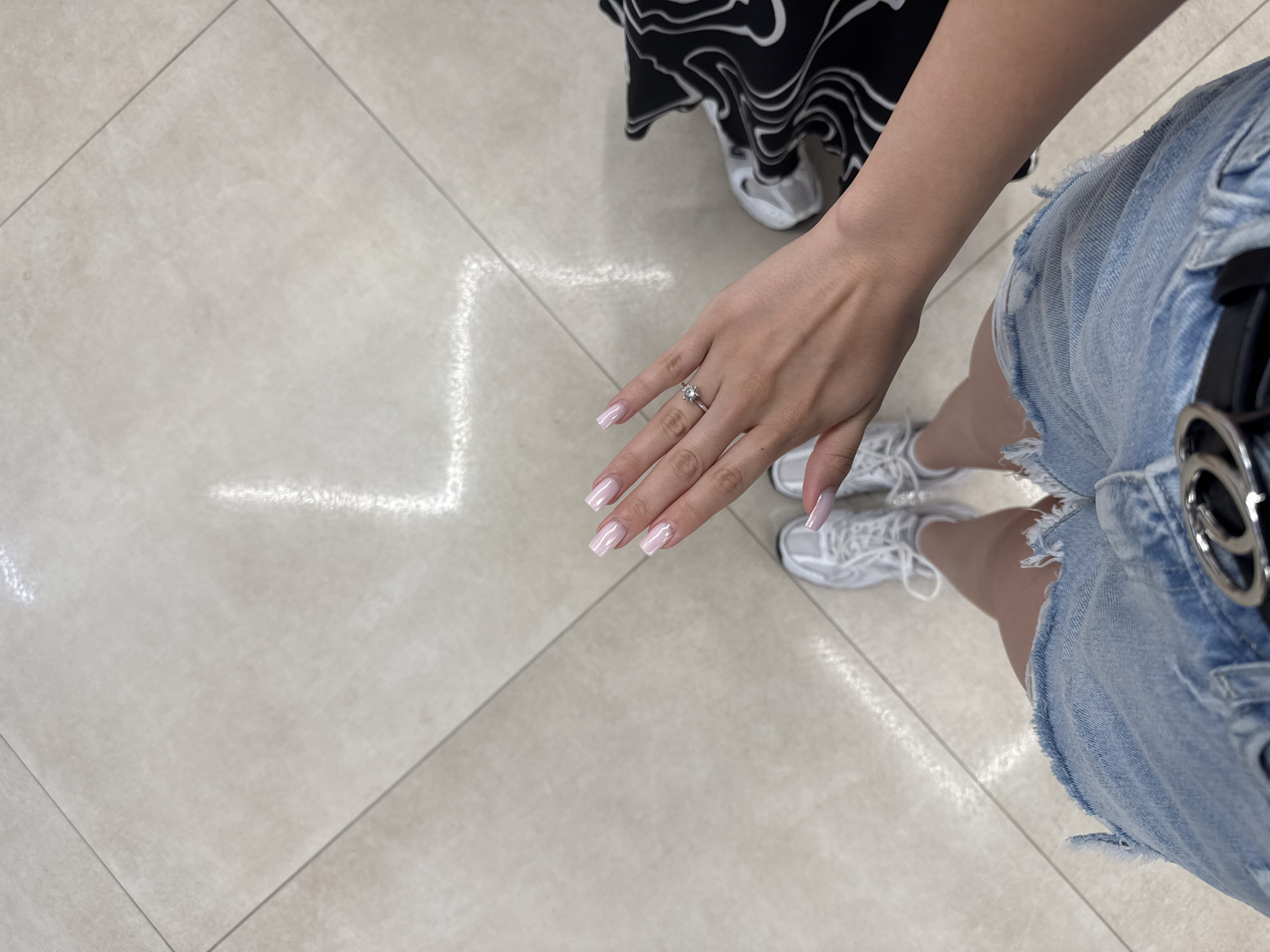 The image shows a person's legs, hand, and a portion of their torso, viewed from a high angle, standing on a tiled floor. The floor tiles are light beige with a subtle pattern, and light reflects off them, creating bright rectangular shapes. The person is wearing distressed denim shorts, a black and white patterned garment on their right side, and white sneakers. The person's left hand is extended, displaying manicured nails with a pale pink, iridescent polish and a ring with a central diamond on the ring finger. The scene appears to be indoors, possibly a commercial space or a well-lit hallway, given the floor tiling and lighting. No specific landmarks or indicators place the location in Chișinău, Moldova.