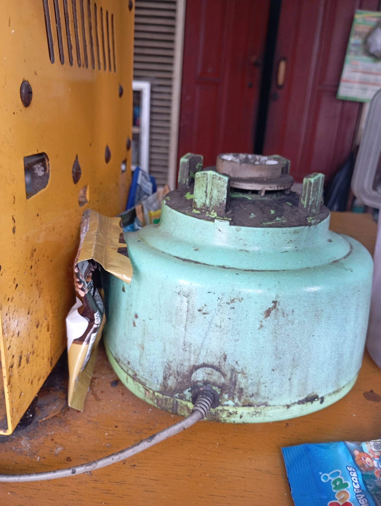 The image shows a close-up of a dusty, pale green blender base on a wooden surface, with a yellow appliance to the left. The blender base appears old and worn, with dirt and debris accumulated around its top and sides. A power cord extends from the bottom of the blender. To the left, a portion of a yellow appliance with ventilation slits is visible. A crumpled snack wrapper is caught between the yellow appliance and the blender base. In the background, a dark red door or wall is partially visible, along with some indistinct objects. The scene appears to be an indoor setting, possibly a storage area or a workshop, in Babelan, Indonesia.
