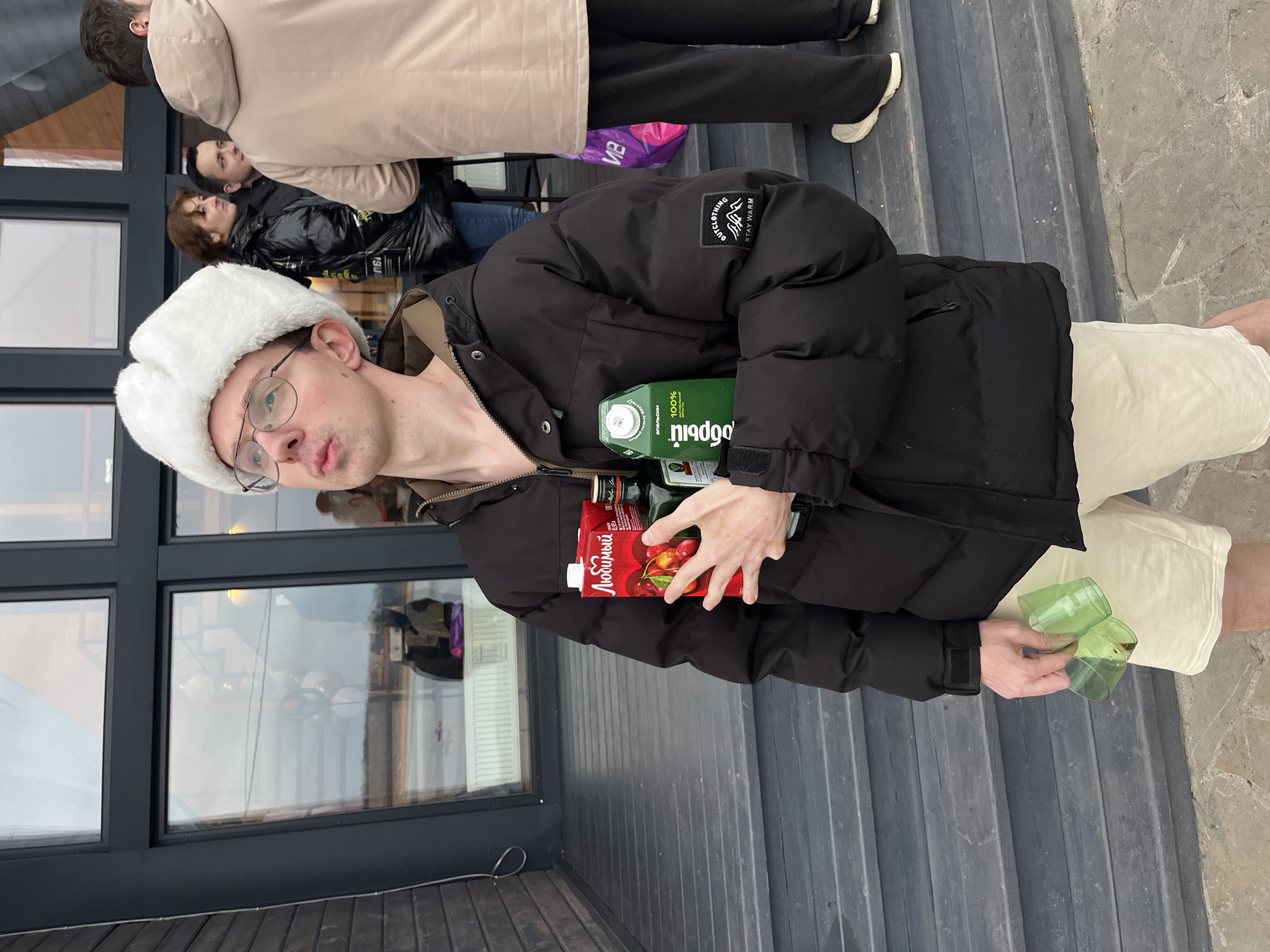 A man wearing a white fur hat and glasses is standing outside a building in Borshchevo, Russia. He is holding a carton of "Любимый" cherry juice, a carton of "Я" juice, and two green plastic cups. He's wearing a black winter coat and beige shorts. Other people are standing behind him. The building has large glass windows.
