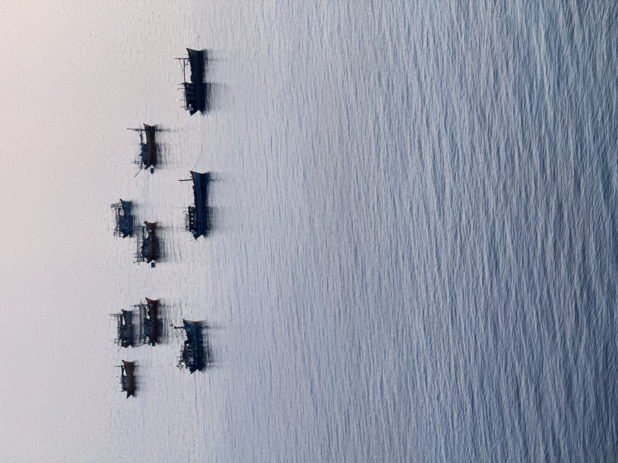 The image shows a serene seascape with several fishing boats anchored in calm waters. The boats, varying in size and color, are clustered together in the upper portion of the frame, leaving the majority of the image to a vast expanse of water that transitions from a light, hazy sky to a deeper blue towards the foreground. The water exhibits gentle ripples, creating a textured surface. The sky appears to be in a soft, diffused light, possibly dawn or dusk, contributing to the tranquil atmosphere. The setting is likely the coastal waters around Lý Sơn Special Administrative Region, Vietnam, a region known for its fishing industry and picturesque seascapes.