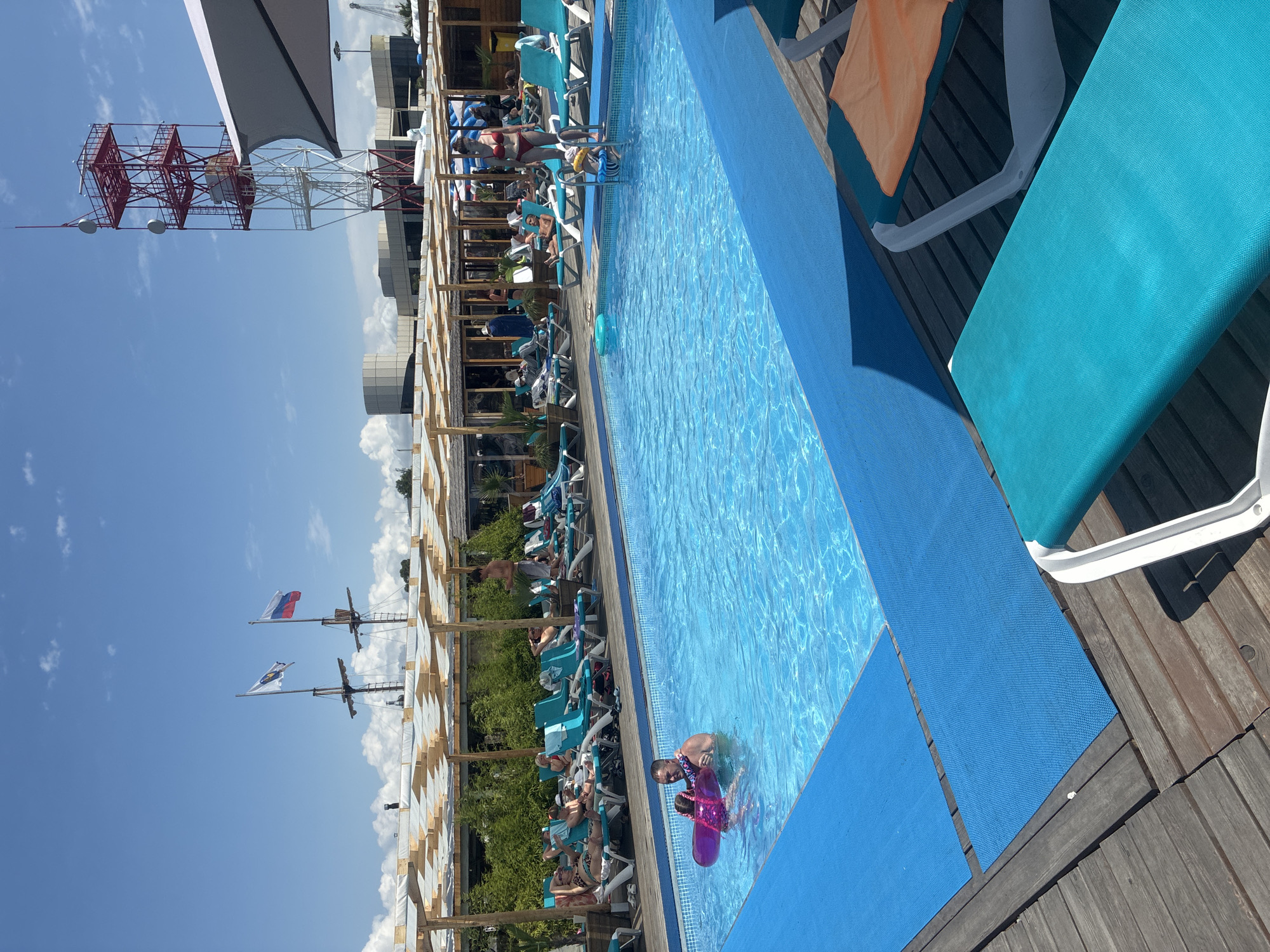 The image depicts an outdoor swimming pool area on a sunny day, likely in Sirius, Russia, given the presence of a Russian flag on a ship's mast in the background. The pool is a vibrant blue with a blue matting surrounding it on a wooden deck. Several lounge chairs, some occupied by sunbathers, are arranged around the pool. In the foreground, a man is holding a young girl in the water; the girl is wearing a pink inflatable ring. Further back, a woman in a red bikini stands near the pool steps. Other people are visible lounging on chairs and walking around the area. A distinctive red and white radio tower stands tall in the background, along with several buildings and a portion of what appears to be a ship. The sky is clear blue with scattered white clouds.