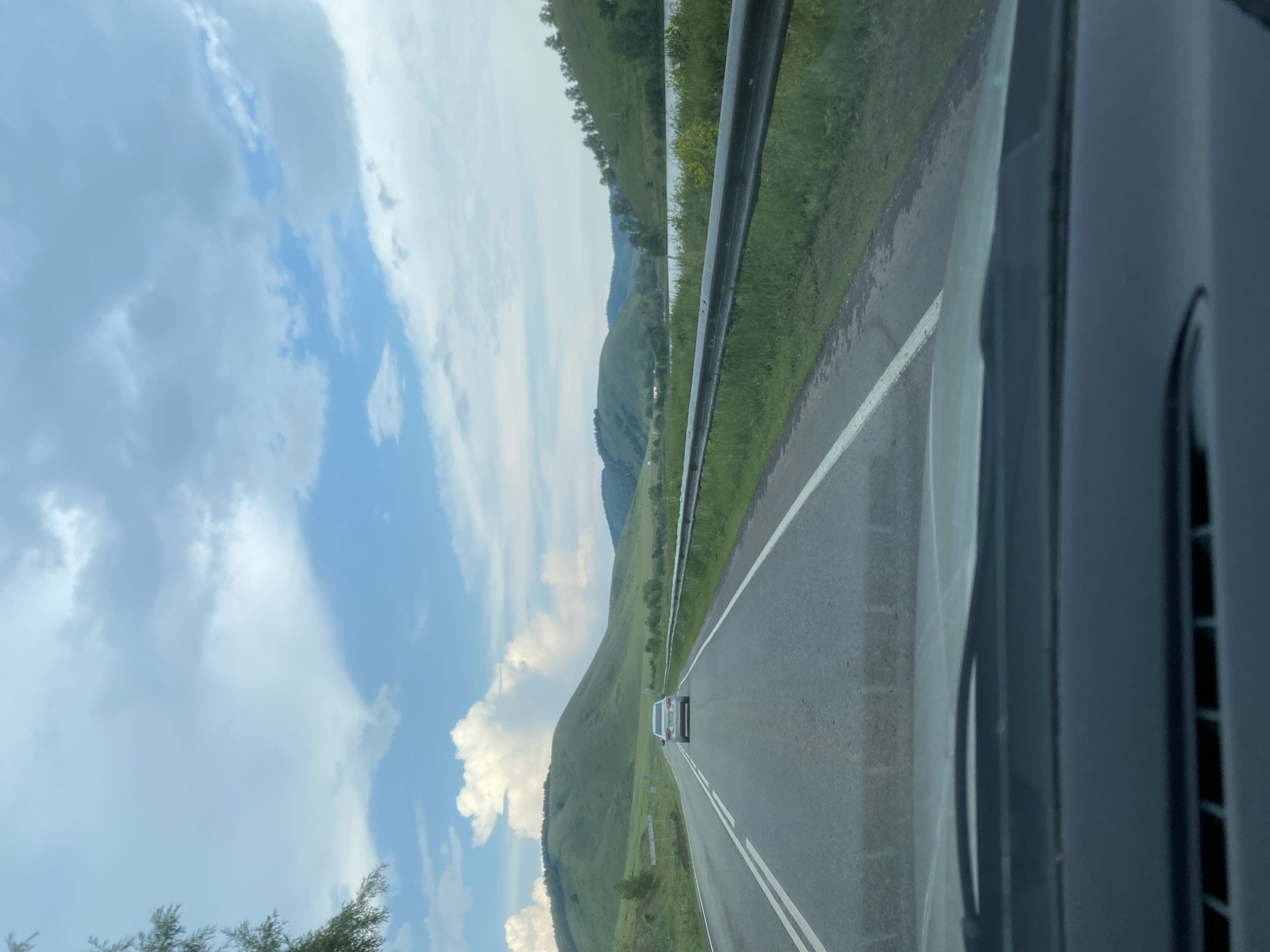 The image captures a scenic view of a two-lane road in the city of Kugarchi, Russia, with a silver sedan driving ahead. The road is flanked by lush green grassy shoulders and a metal guardrail on the right side, separating the road from a body of water and more greenery. Rolling green hills dominate the background, with a few scattered trees. The sky is a mix of blue and white clouds, with a large, fluffy cloud formation over one of the hills. The photograph appears to be taken from a moving vehicle, indicated by the perspective and the blurred foreground of a car's dashboard. The environment suggests a rural or semi-rural setting within Kugarchi, Russia, characterized by natural landscapes and a clear sky.