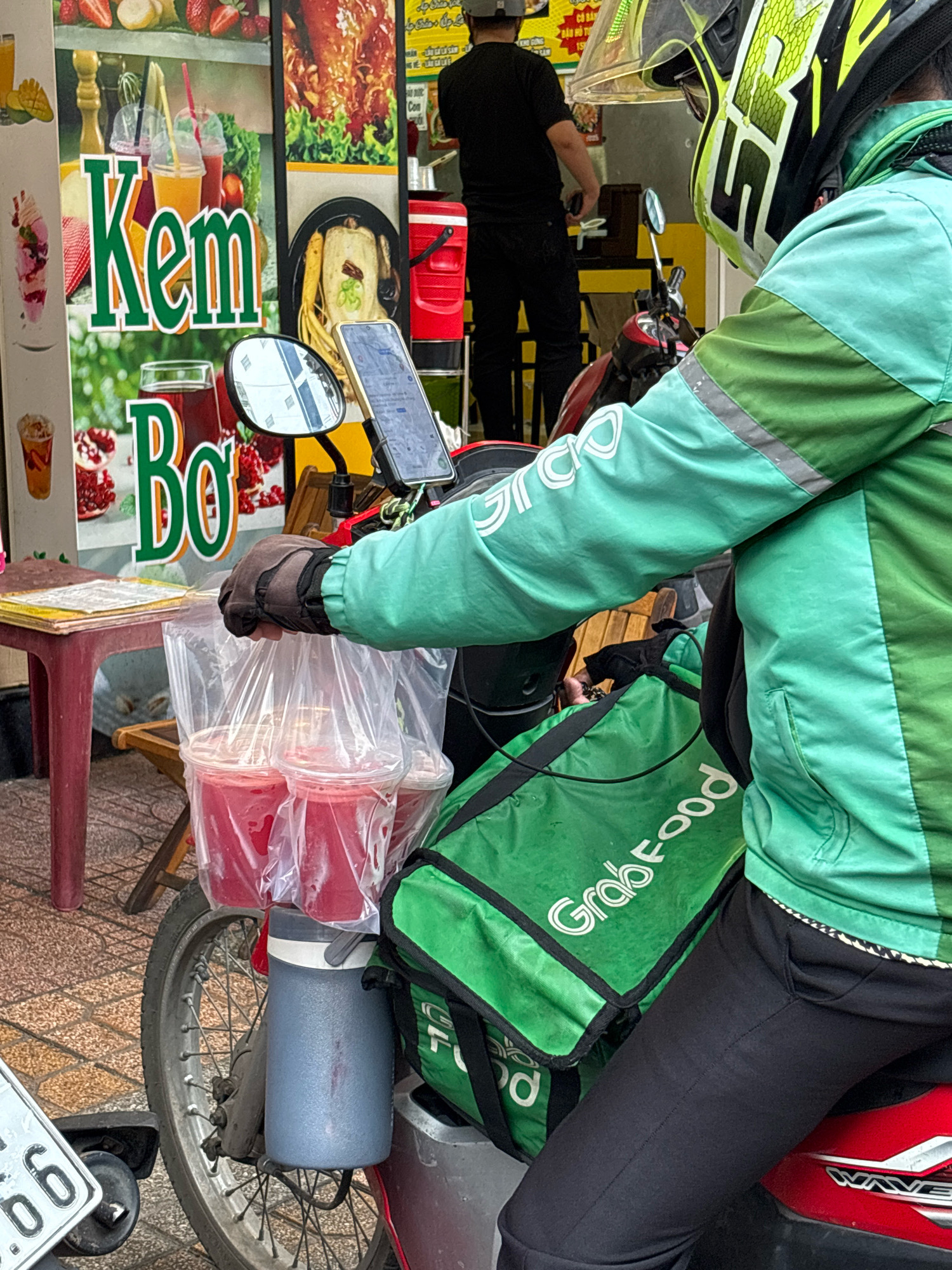 The image captures a street scene in Nha Trang, Vietnam, focusing on a food delivery person on a scooter. The delivery person, wearing a teal and green uniform with a white "GrabFood" logo on their jacket and a neon yellow and black helmet, is holding a plastic bag containing three cups of red beverage. The scooter has a large green insulated bag labeled "GrabFood" behind the driver, and a grey water bottle holder attached to the frame. In the background, a stall displays various food items, including fruits, cooked poultry, and drinks, with Vietnamese text visible on signage. The setting appears to be a busy urban environment with tiled sidewalks.