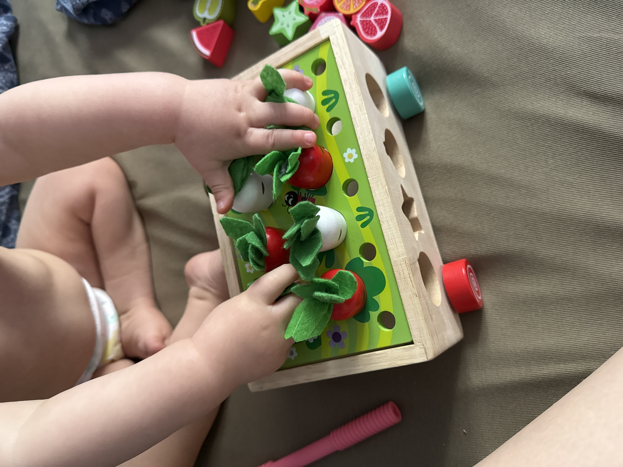 A child, wearing a diaper, is seated on an olive-colored surface and interacting with a wooden toy box. The box is green with illustrations of plants and features holes where toy radishes and turnips can be inserted and "pulled" from the box. The child's hands are visible as they manipulate the toys. Scattered around the toy box are wooden fruits and vegetables. A pink toy sits near the bottom of the image. The image provides no information to suggest the location is Moscow, Russia.