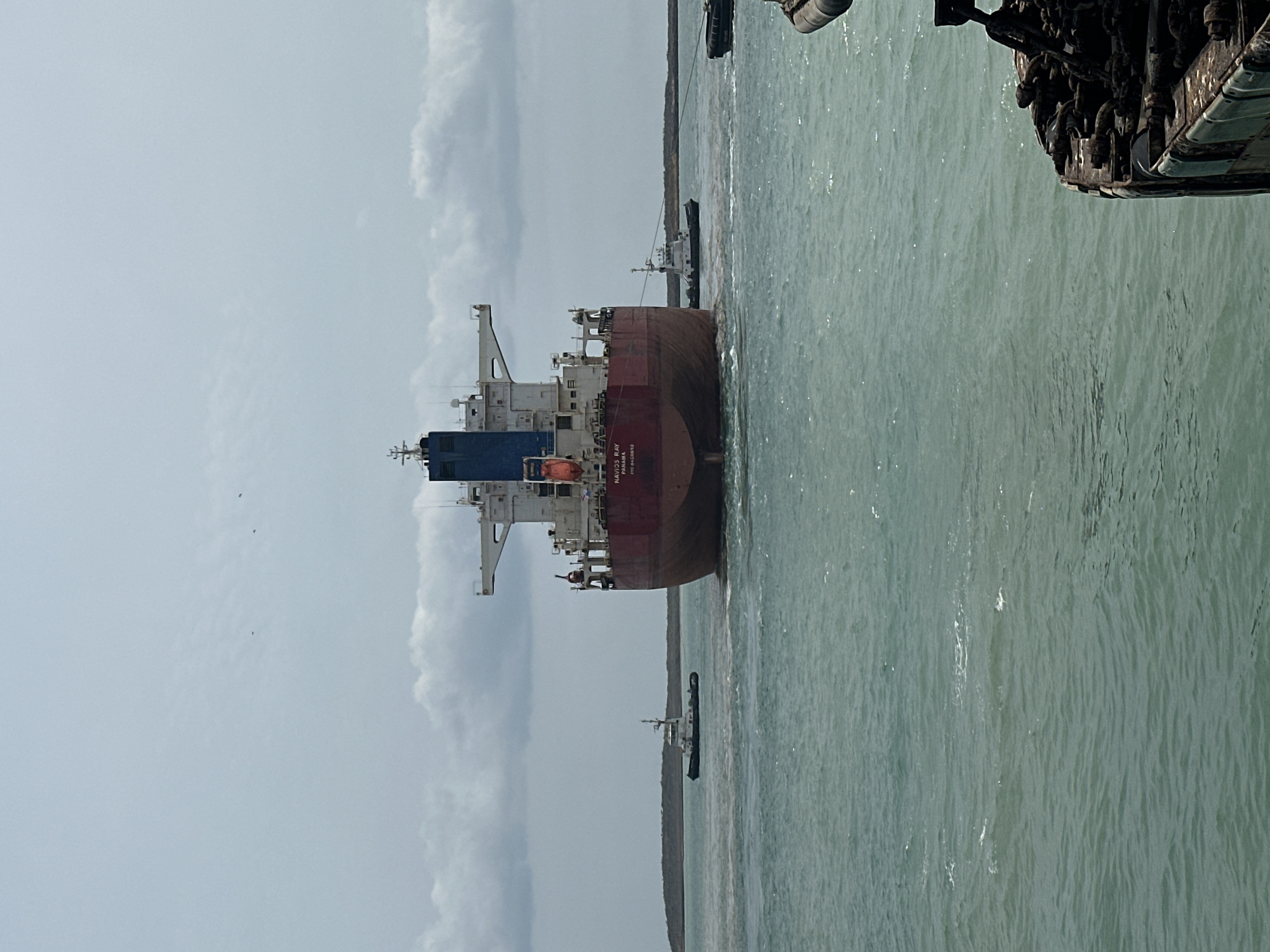 The image displays a large bulk carrier, identified as the "NAVIOS RAY" (IMO 9405512) and flagged in Panama, being maneuvered by two tugboats in the coastal waters near Uribia, Colombia. The bulk carrier is viewed from its stern, revealing a reddish-brown hull, a white mid-deck, and a blue upper deck structure with a mast. Two white tugboats, each with a dark hull stripe, are actively assisting the main vessel; one is positioned to the port side, and another is to the starboard side, connected to the "NAVIOS RAY" by a towing line. The water is a teal-green with slight surface ripples and localized turbulence around the moving vessels. In the background, a low-lying, sparsely vegetated landmass forms the horizon line. The sky is overcast with a blanket of grey and white clouds. In the lower right corner, a dark, weathered structure with chains and fittings, indicative of another vessel's deck or a pier, frames the foreground. No individuals are clearly discernible.