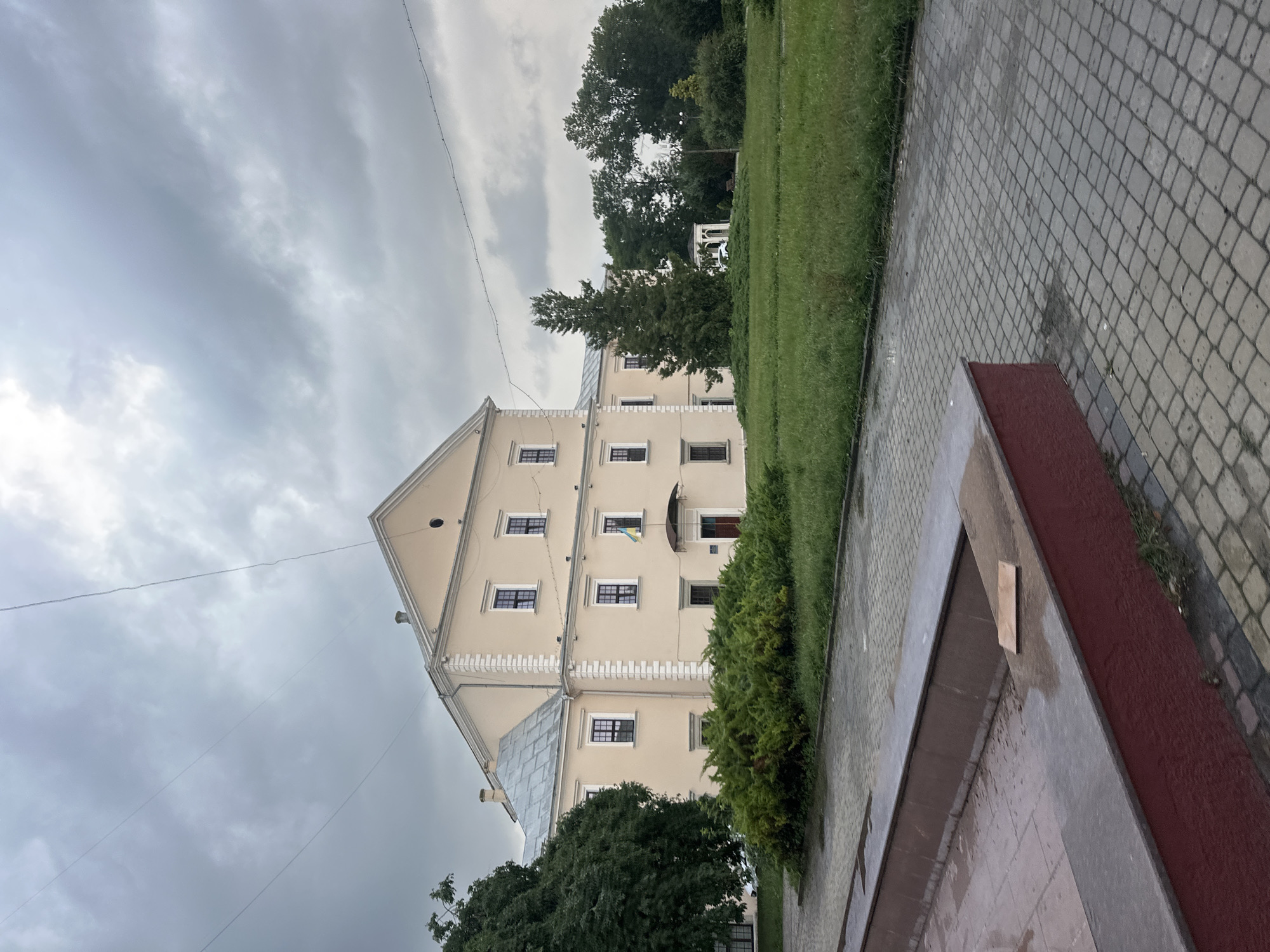 The image shows the Ternopil Regional Military History Museum (also known as Ternopil Castle) in Ternopil, Ukraine. The castle is a large, pale yellow building with a grey roof and dark-framed windows. A Ukrainian flag is visible hanging from the building. In front of the castle, there is a green lawn with trees and bushes. A paved walkway with red-trimmed planters is in the foreground. The sky is cloudy and overcast, suggesting a cool, possibly damp day. There are no people visible in the image.