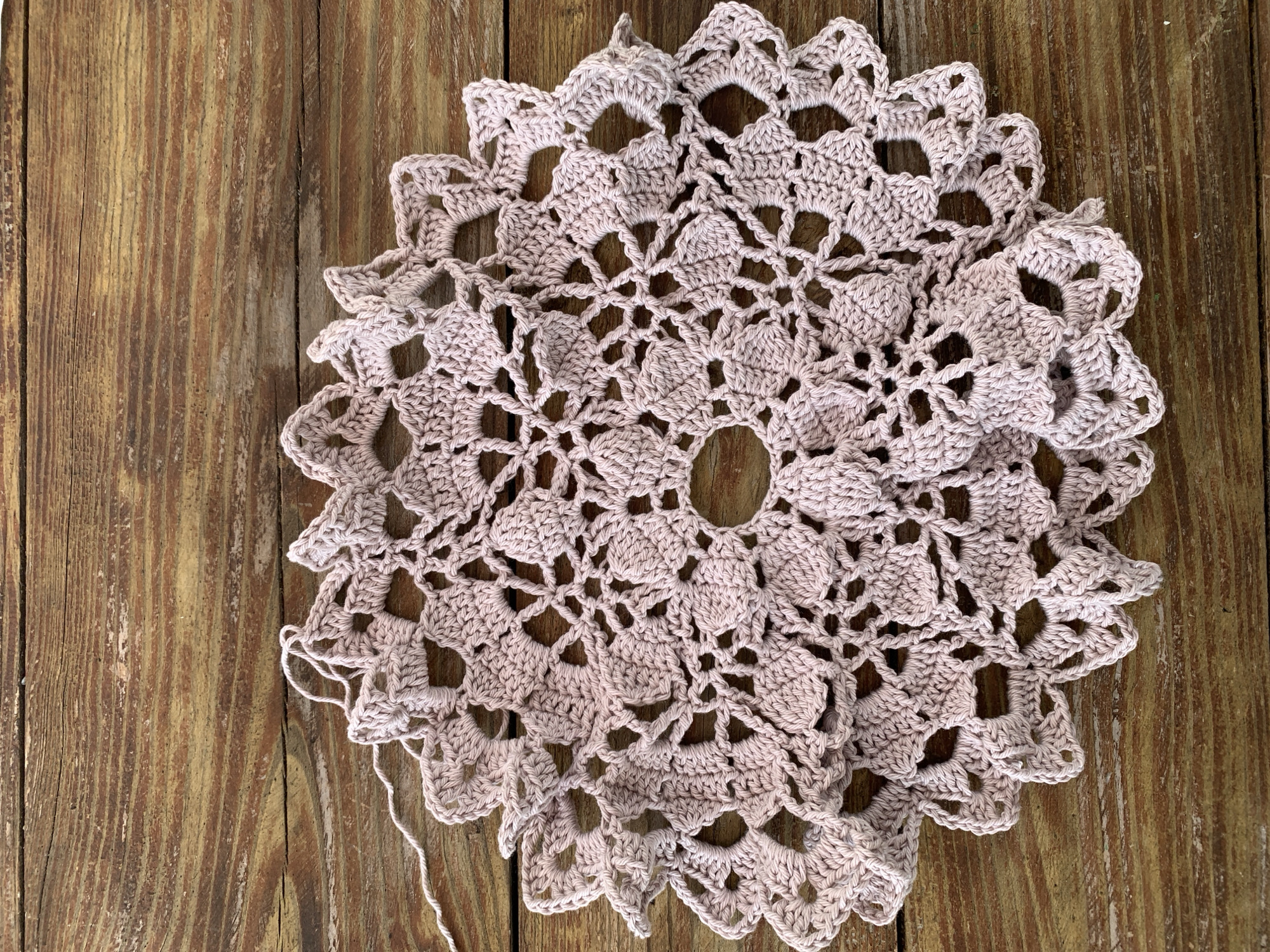 A light pink, crocheted doily is partially folded and lies on a weathered, wooden surface. The doily has a circular, intricate pattern with open spaces and decorative elements that resemble petals or leaves. A loose white thread extends from the left edge of the doily. The wooden surface consists of planks with visible grain and a distressed finish, suggesting outdoor or rustic placement. No people or animals are present. The scene appears to be a close-up, overhead shot. The location provided, Капцёўскі сельскі Савет, Belarus, is not visually represented in the image itself.