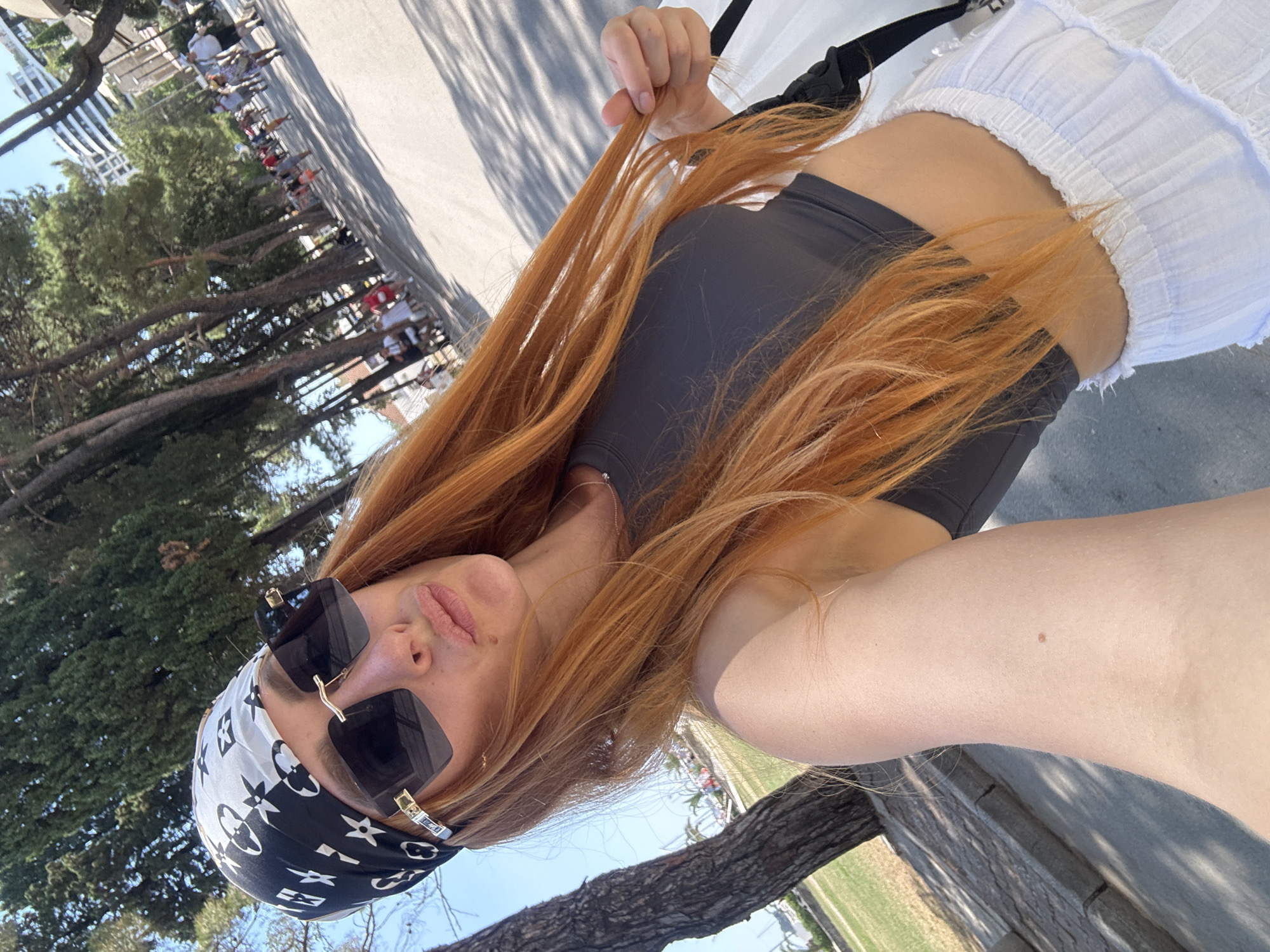 The image shows a young woman with long, reddish-blonde hair, wearing a dark grey sports bra and white ruffled shorts. She has a black and white bandana with a repeating logo pattern tied around her head, and large, dark sunglasses. She is taking a selfie outdoors on a sunny day, possibly in the late morning or early afternoon, given the bright sunlight and clear sky.
The background includes trees with sparse foliage, suggesting a warm season like spring or summer. In the distance, there are buildings and a group of people walking in a line, some appearing to be seated or on display, which could indicate a public event or a promenade. The ground is paved, and there are shadows cast by trees and possibly a railing. A tree trunk is visible in the foreground, partially obscuring the view. The woman appears relaxed and is holding a strand of her hair. The setting evokes a casual, outdoor atmosphere in a Mediterranean location like Budva, Montenegro, given the context. No legible text is visible in the image.