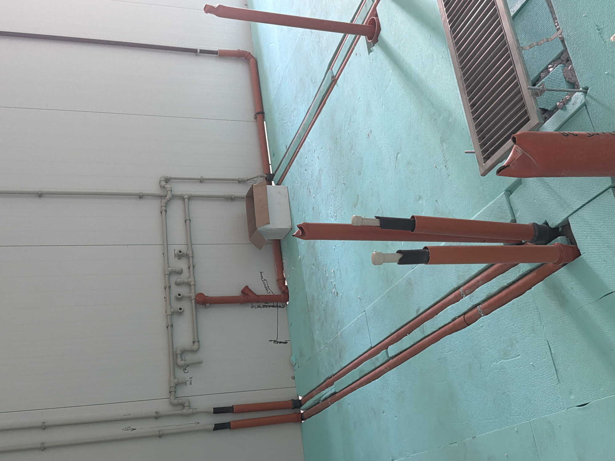 This is an interior photograph of a room under construction, likely in Nikolaevo, Bulgaria. The room has light-colored, paneled walls and a light blue, foam-insulated floor. The floor is covered with what appear to be large, interlocking foam tiles.
The primary focus of the image is the extensive network of orange and grey pipes and conduits installed on the walls and floor. On the wall, a series of grey pipes with various T-junctions and connectors are visible, some with handwritten markings. A prominent red pipe emerges from the floor and connects to this network. Several other vertical orange pipes emerge from the floor, some capped with black material and others with grey plastic caps. One of these vertical pipes has a label that appears to read "стерилизатор" (sterilizer in Cyrillic).
In the foreground, several orange pipes are laid out on the blue floor, some running parallel to each other in channels cut into the foam insulation. A stainless steel grate, possibly a floor drain or ventilation cover, is positioned on the floor towards the right side of the image. A cardboard box is visible near the wall on the left. The overall impression is of an industrial or utility space where plumbing and electrical systems are being installed. There are no people or animals present in the image.
