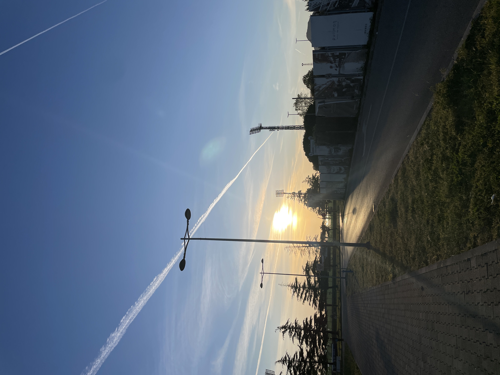 The image shows a street scene in Bar, Montenegro, illuminated by the rising sun. A paved path runs along the bottom right of the frame, with grass and trees separating it from an adjacent road. Large banners or billboards stand along the road edge. In the sky, contrails from airplanes are visible against a clear blue sky. Streetlights are silhouetted against the sun, and floodlights can be seen in the distance. The Pullman Hotel sign is visible on the banners.