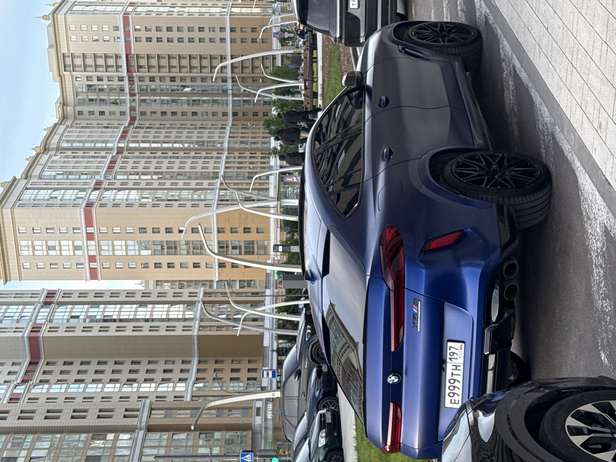 The image shows a row of parked cars on a street in Moscow, Russia. The closest car is a blue BMW X6M, with the license plate E999TH197 RUS. Tall apartment buildings are visible in the background. There are people walking on a sidewalk in the distance. The weather appears to be overcast.