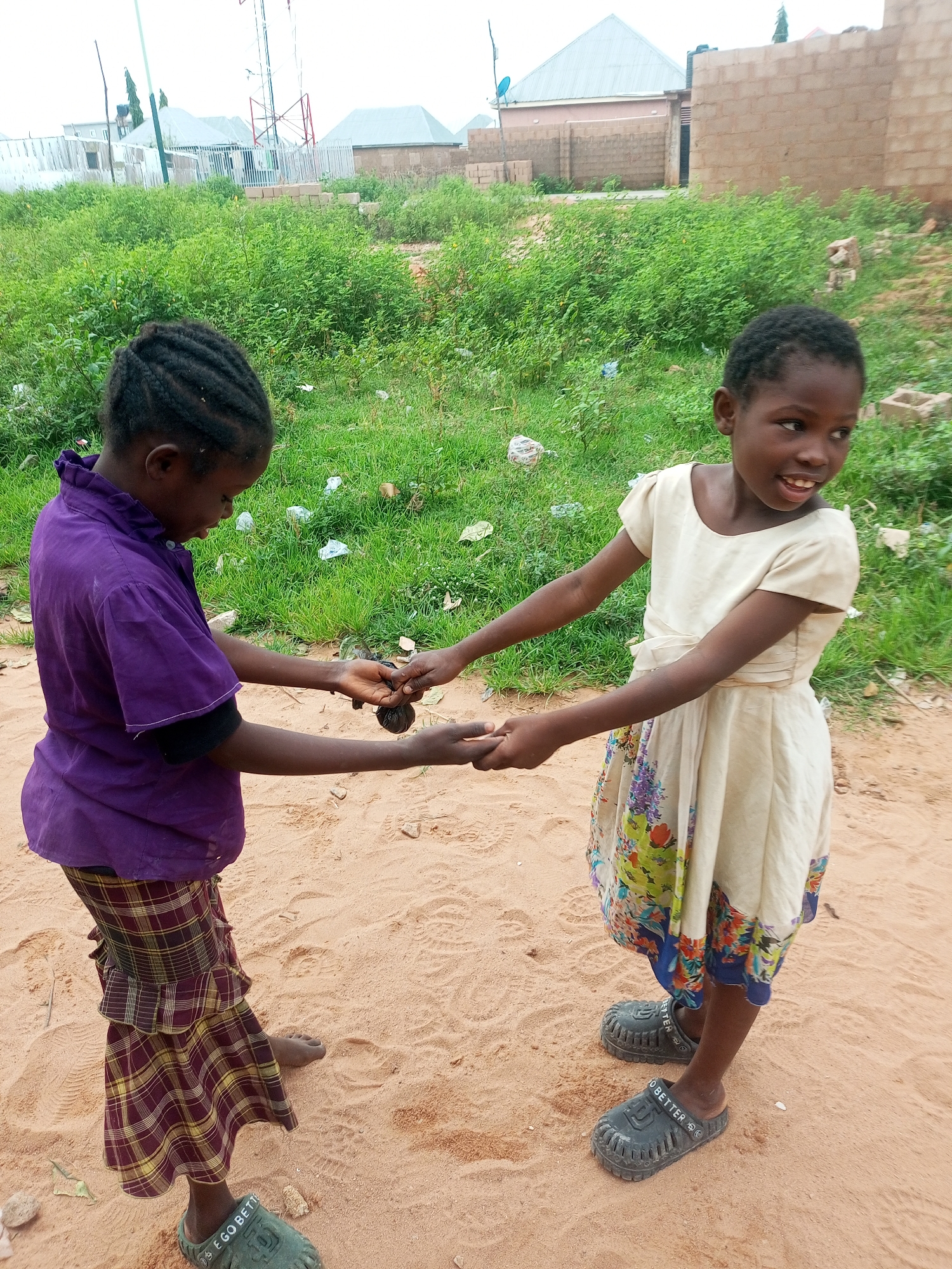 Two young girls are engaged in a playful interaction on a sandy ground with sparse vegetation and buildings in the background. The girl on the left, wearing a purple shirt and a plaid skirt, is holding her hands out, seemingly offering or exchanging an object with the girl on the right. The girl on the right is wearing a light-colored dress with a floral pattern and is smiling, looking to her right. Both girls are barefoot or wearing open-toed footwear. The environment appears to be an outdoor space in a somewhat undeveloped area, possibly on the outskirts of Tempure, Nigeria, with unfinished brick buildings and some overgrown green plants. The overall scene suggests a moment of childhood play and interaction in a rural or semi-urban setting.