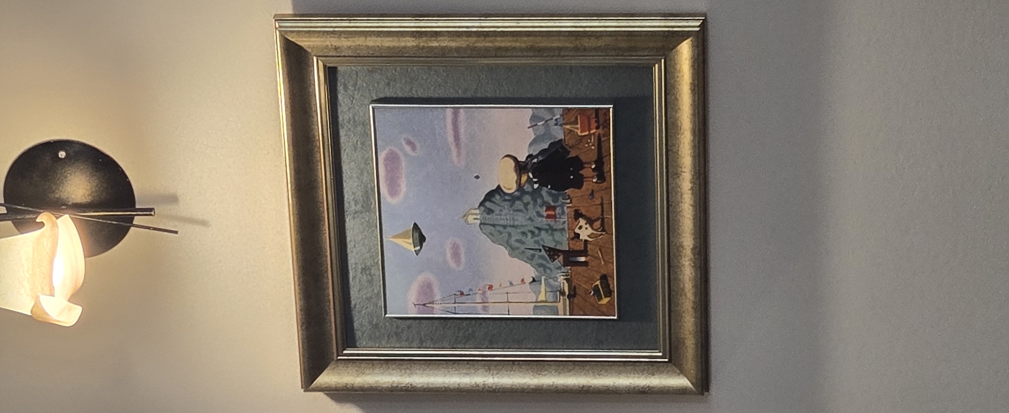The image presents a framed painting hanging on a textured, light-colored wall, illuminated by an adjacent wall-mounted light fixture.
The painting itself depicts a surreal and whimsical scene. At the center is a stylized figure, seemingly a man from the torso up, with a smooth, light-colored, featureless head. He wears a patterned blue-grey jacket and stands on a brown and orange-toned surface resembling a pier or boardwalk. To his right, another figure, dressed in black with a wide, dark hat, is positioned, also with an obscured face. A small, fawn-colored, stylized animal, possibly a dog, sits on the pier to the left of the central figure.
In the background, a sailboat with colorful pennant flags strung on its rigging is visible. A small, golden, pyramid-shaped object appears to float in the lavender-hued sky, which is dotted with abstract, blotchy purple shapes. The scene evokes a quiet, contemplative, and slightly mysterious atmosphere, typical of surrealist art.
The artwork is set within a golden, ornate frame that features a grey-green inner matte.
To the left of the framed painting, a modern wall sconce is affixed. It has a dark, circular base and thin, radiating metal spokes. A curved, translucent shade, shaped somewhat like a flame, emits a warm, yellow-orange light, casting a glow on the wall and parts of the painting. The wall itself is a uniform, light beige or off-white. No visible text is present in the media file. The maritime theme of the painting, with a pier and boat, aligns with the coastal context of Budva, Montenegro.